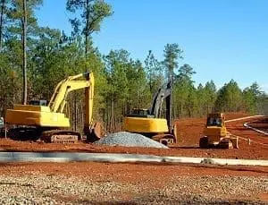 Adherent (formerly Compliance & Risks). Three yellow construction vehicles, two excavators and a smaller loader, on a reddish-brown dirt site with a pile of gravel, a concrete curb, and pine trees in the background.