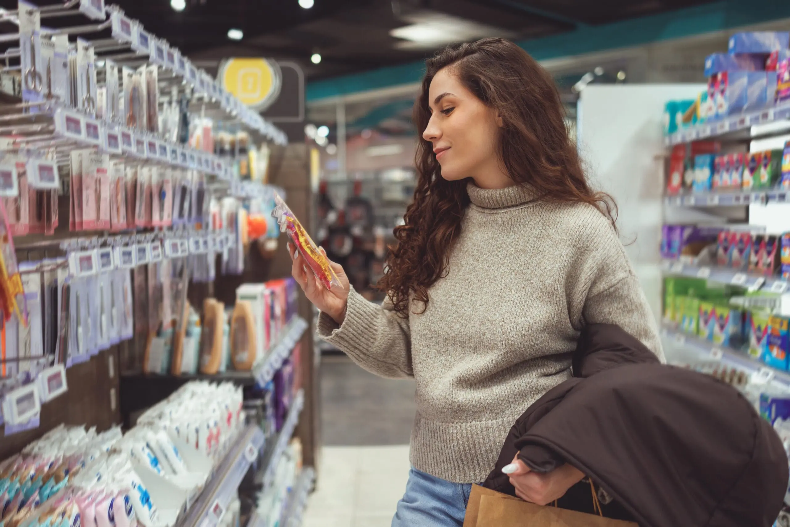 Adherent (formerly Compliance & Risks). A woman in a beige sweater examines a package of colorful hair clips in a store aisle.
