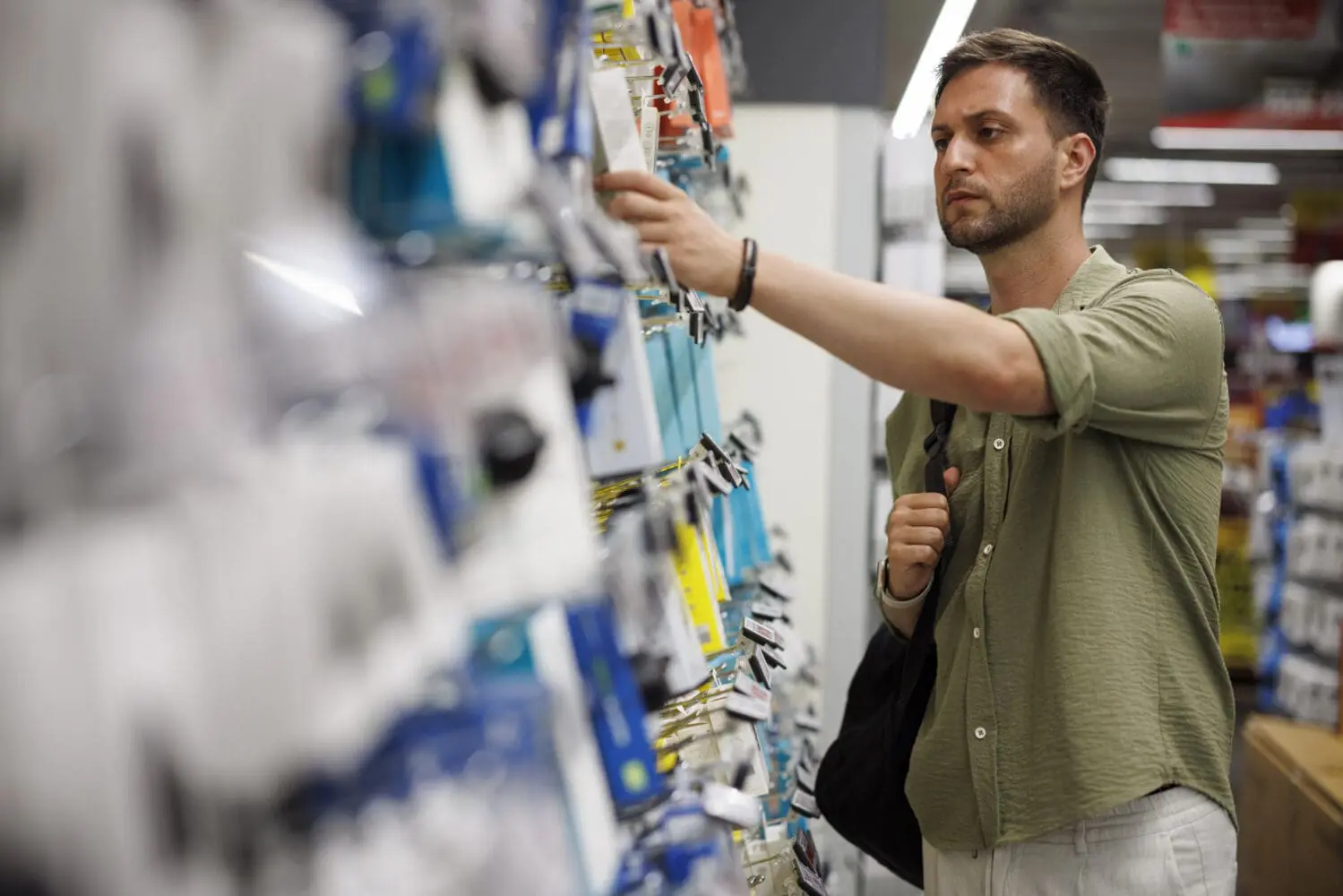 Adherent (formerly Compliance & Risks). A man in a green shirt and light pants reaches for a white-packaged product on a store shelf filled with various items.
