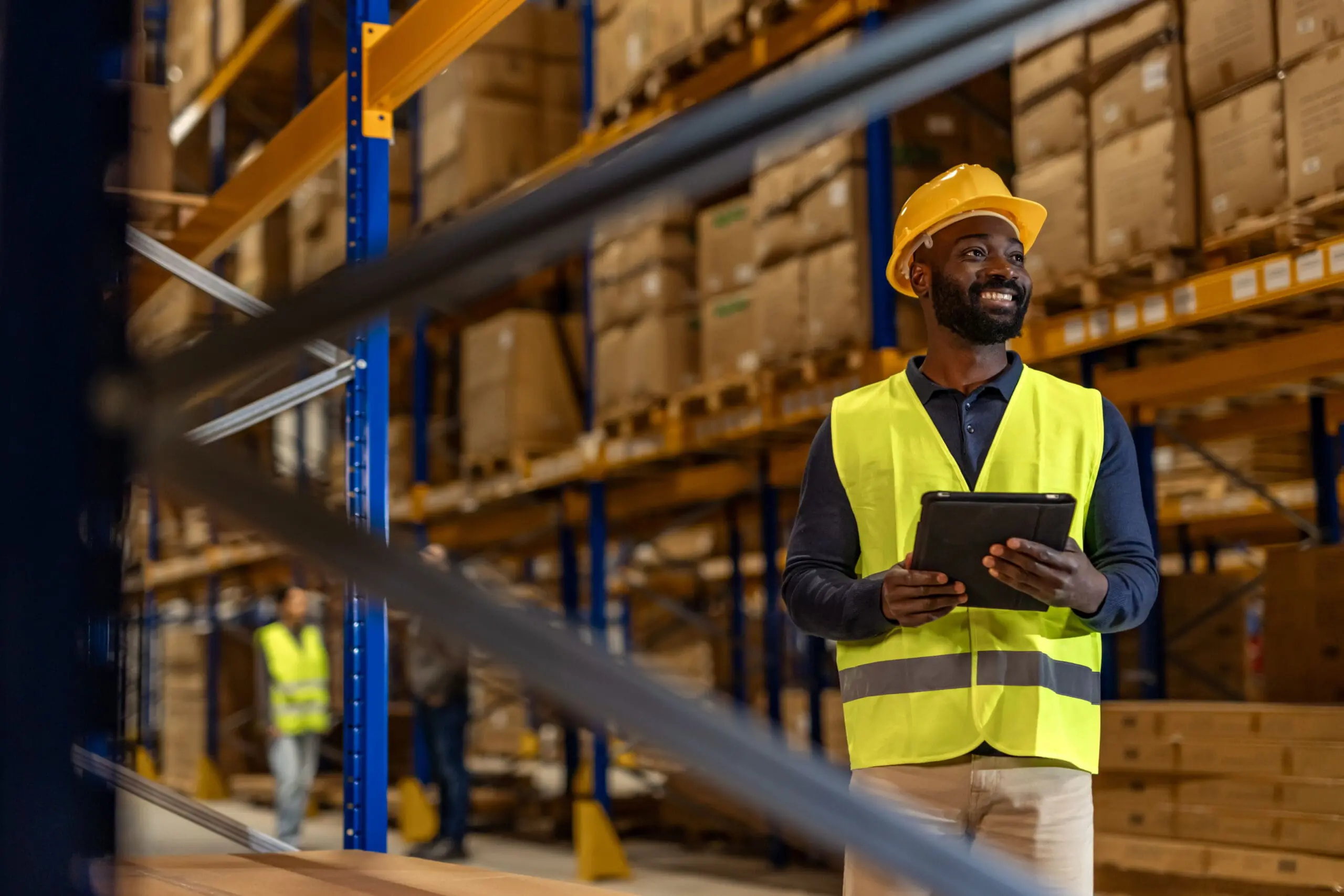Adherent (formerly Compliance & Risks). A smiling Black man in a yellow hard hat and high-visibility vest holds a tablet in a warehouse filled with shelves of boxes.
