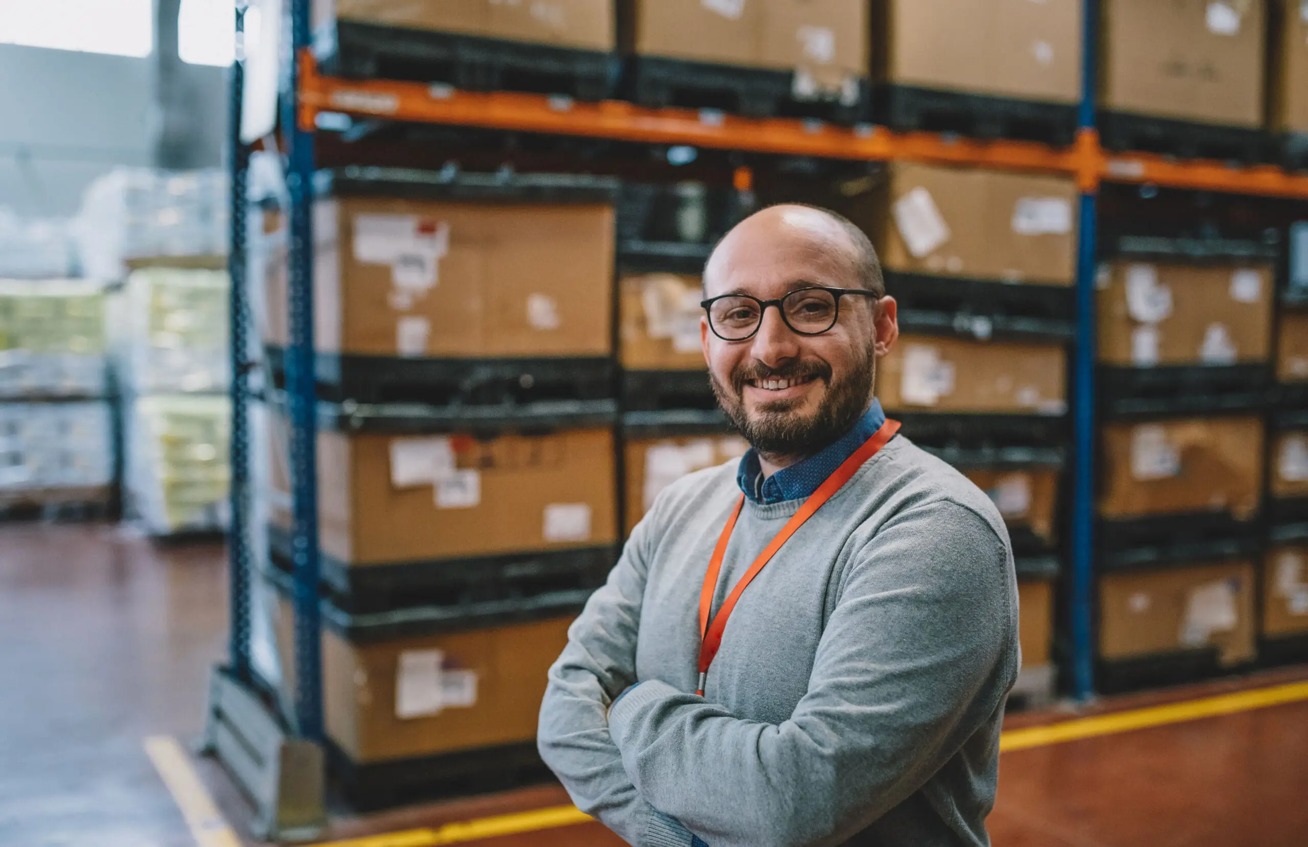 Adherent (formerly Compliance & Risks). A smiling man with glasses and a beard stands with crossed arms in a warehouse filled with cardboard boxes on shelves.