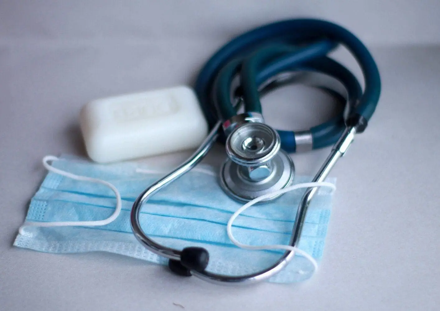 Adherent (formerly Compliance & Risks). A blue stethoscope, a light blue medical face mask, and a white bar of soap on a light gray surface.