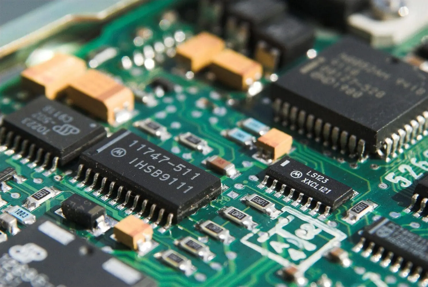 Adherent (formerly Compliance & Risks). Close-up of a green circuit board with various black integrated circuit chips, orange capacitors, and small resistors. Text on chips reads '11747-511 IHSB9111' and 'LS123 XXCL 121'.