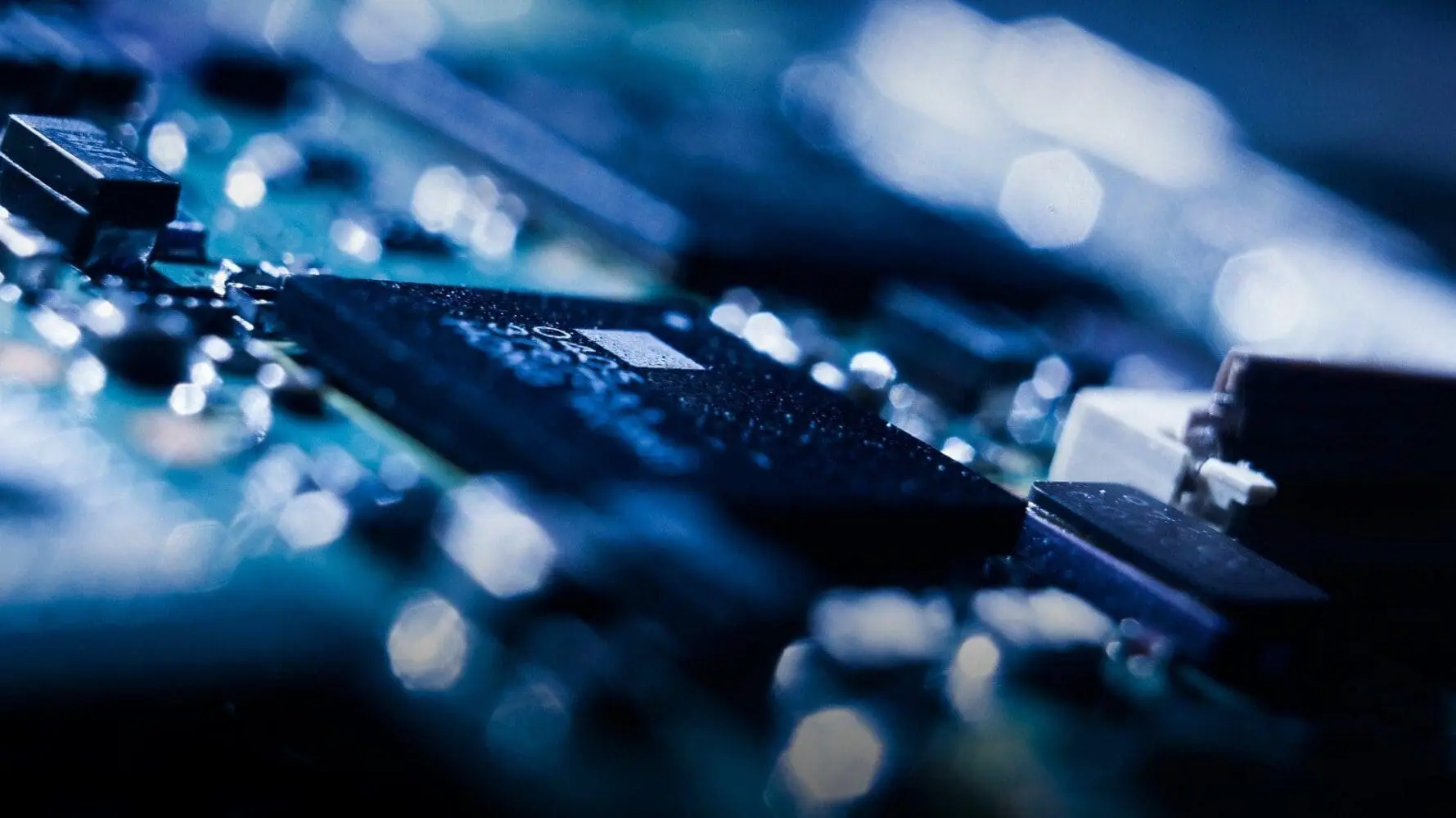 Adherent (formerly Compliance & Risks). Close-up of a circuit board with a dark microchip in focus, surrounded by blurred blue and white lights.