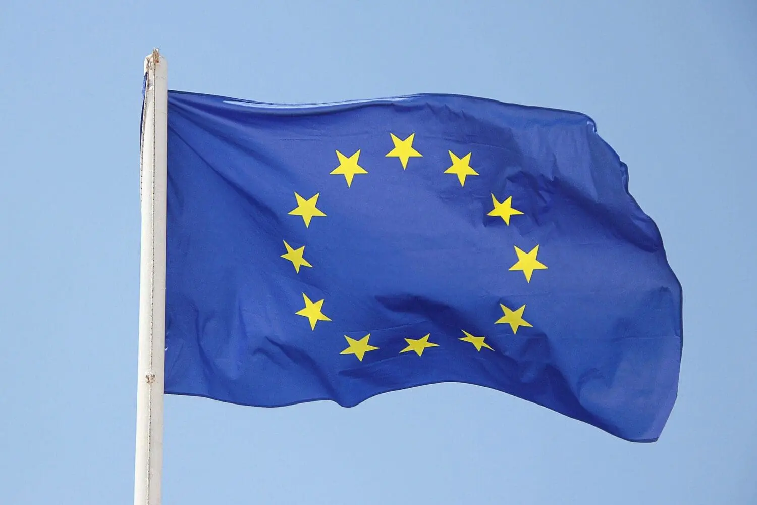 The European Union flag, blue with 12 yellow stars, waving on a white flagpole against a clear blue sky.
