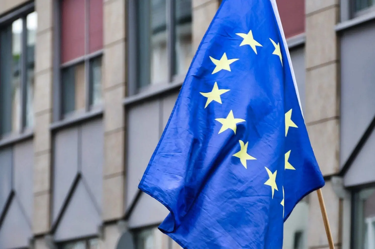 A European Union flag, blue with yellow stars, held by a wooden pole against a blurred building.