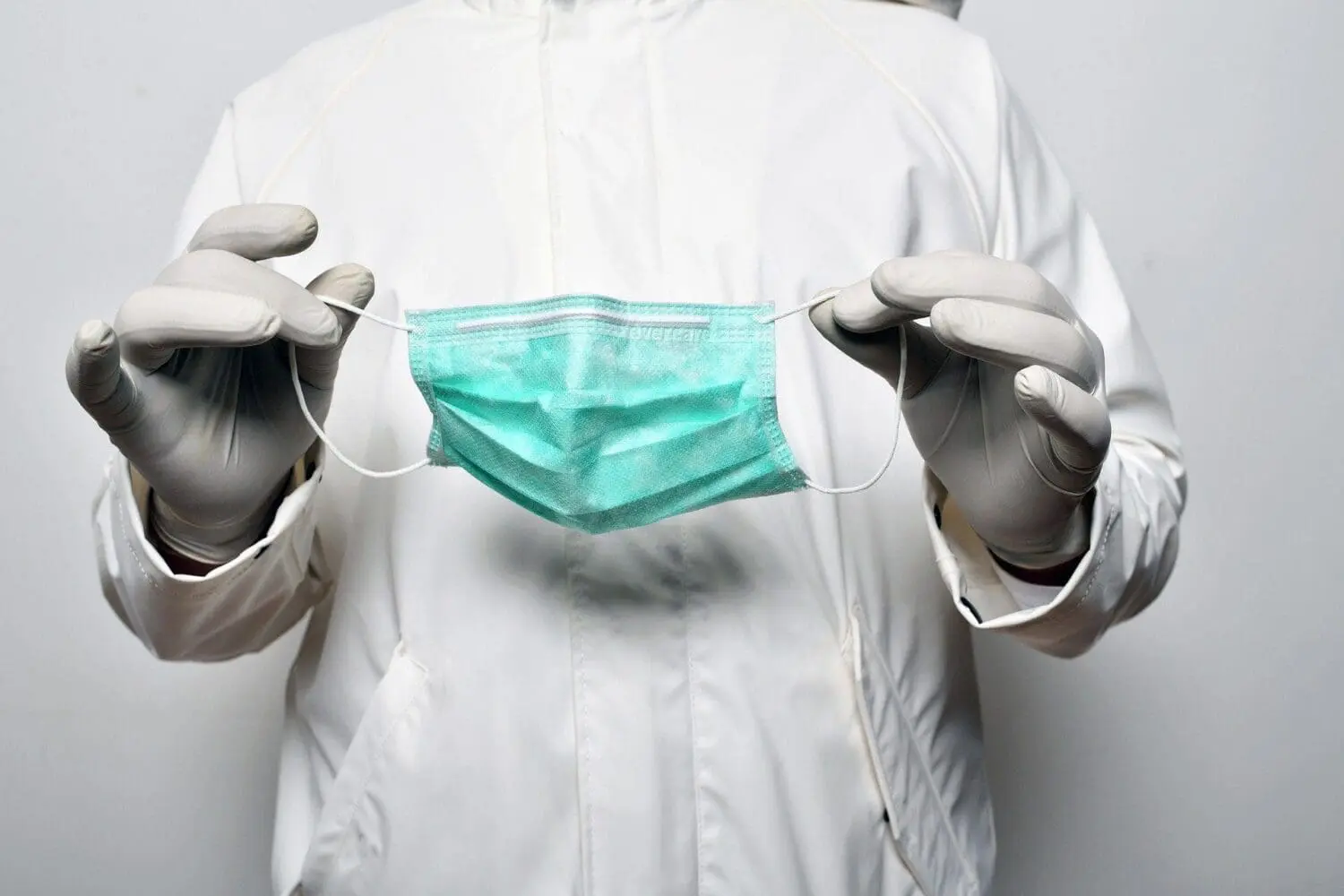 Adherent (formerly Compliance & Risks). A person wearing a white protective suit and grey gloves holds a teal surgical mask by its ear loops.