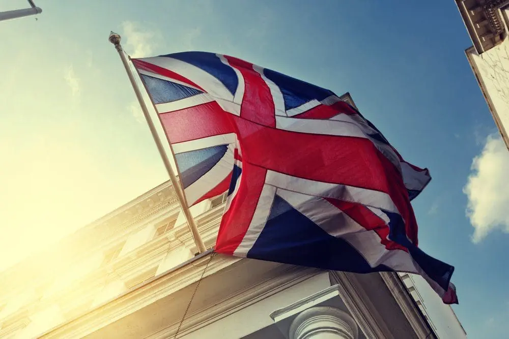 Adherent (formerly Compliance & Risks). A Union Jack flag waves from a flagpole attached to a building, seen from a low angle against a blue sky with a sun flare.