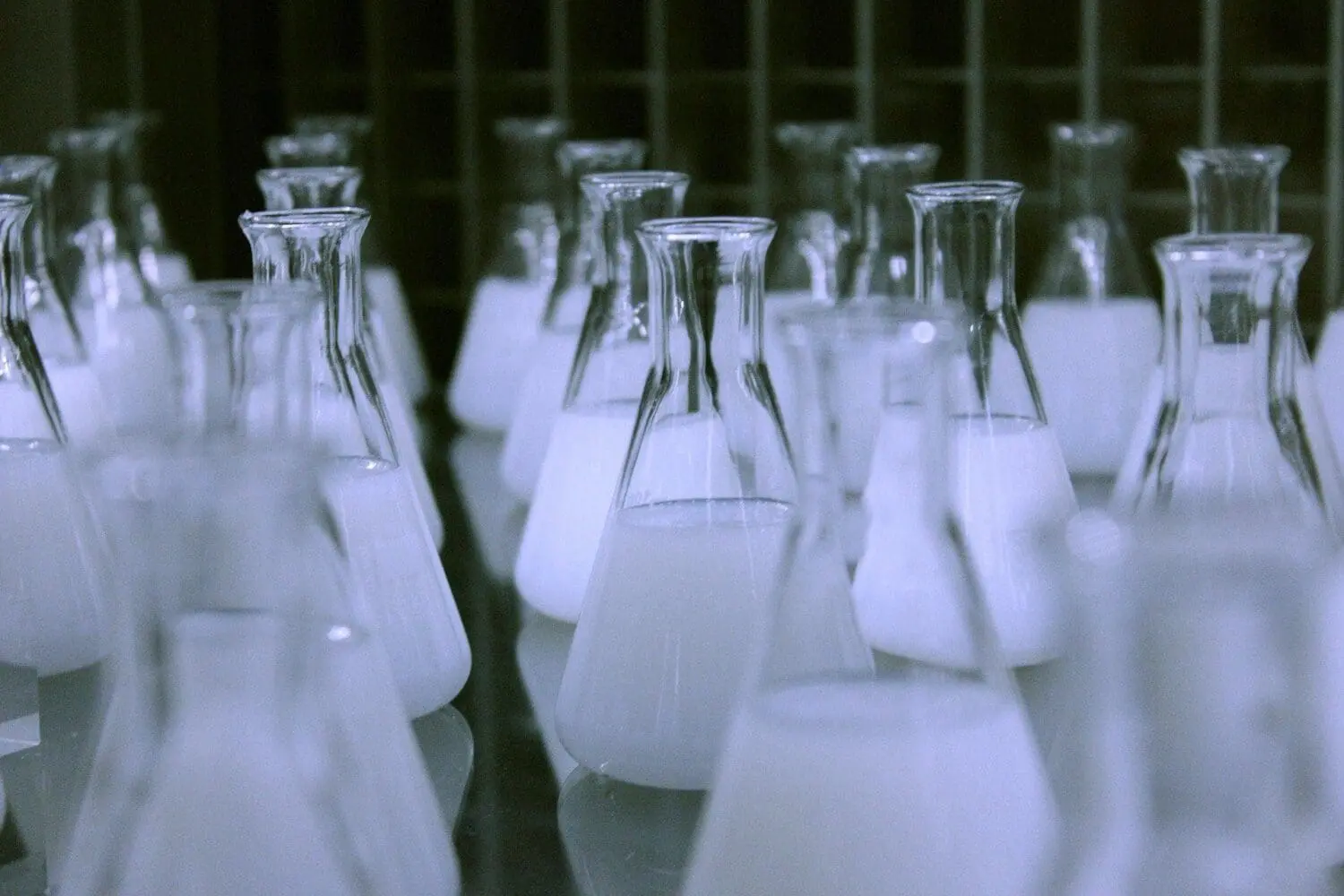 Adherent (formerly Compliance & Risks). Rows of clear glass Erlenmeyer flasks, each containing a white opaque liquid, on a reflective surface in a lab.
