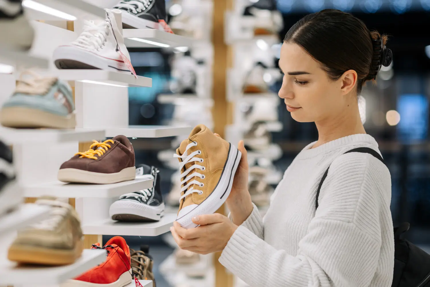 Adherent (formerly Compliance & Risks). A woman in a white sweater holds and looks at a tan high-top sneaker in a shoe store, with other shoes displayed on shelves in the background.