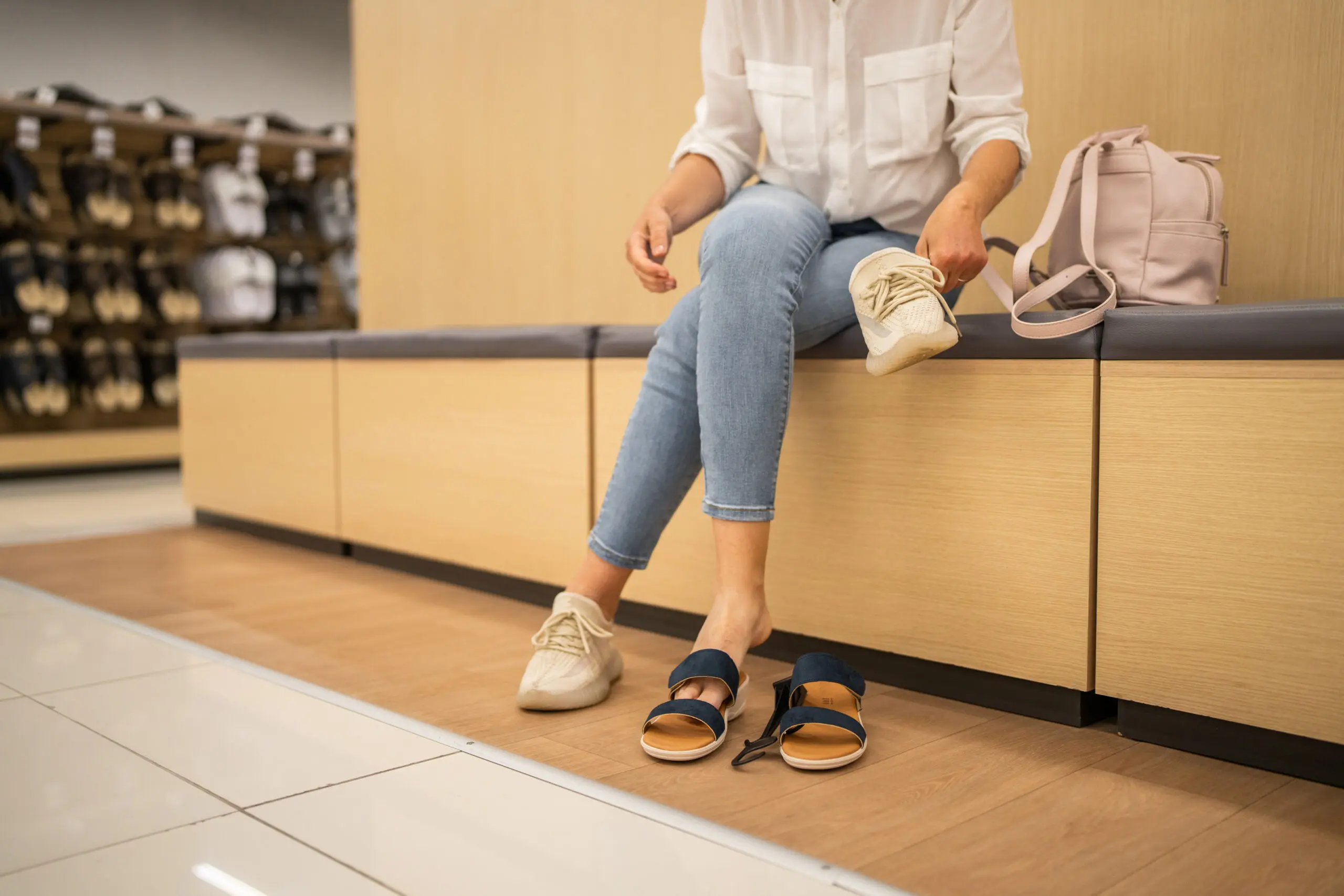 Adherent (formerly Compliance & Risks). A person in a white shirt and blue jeans sits on a bench, holding a beige sneaker, with blue sandals on the floor in a shoe store.