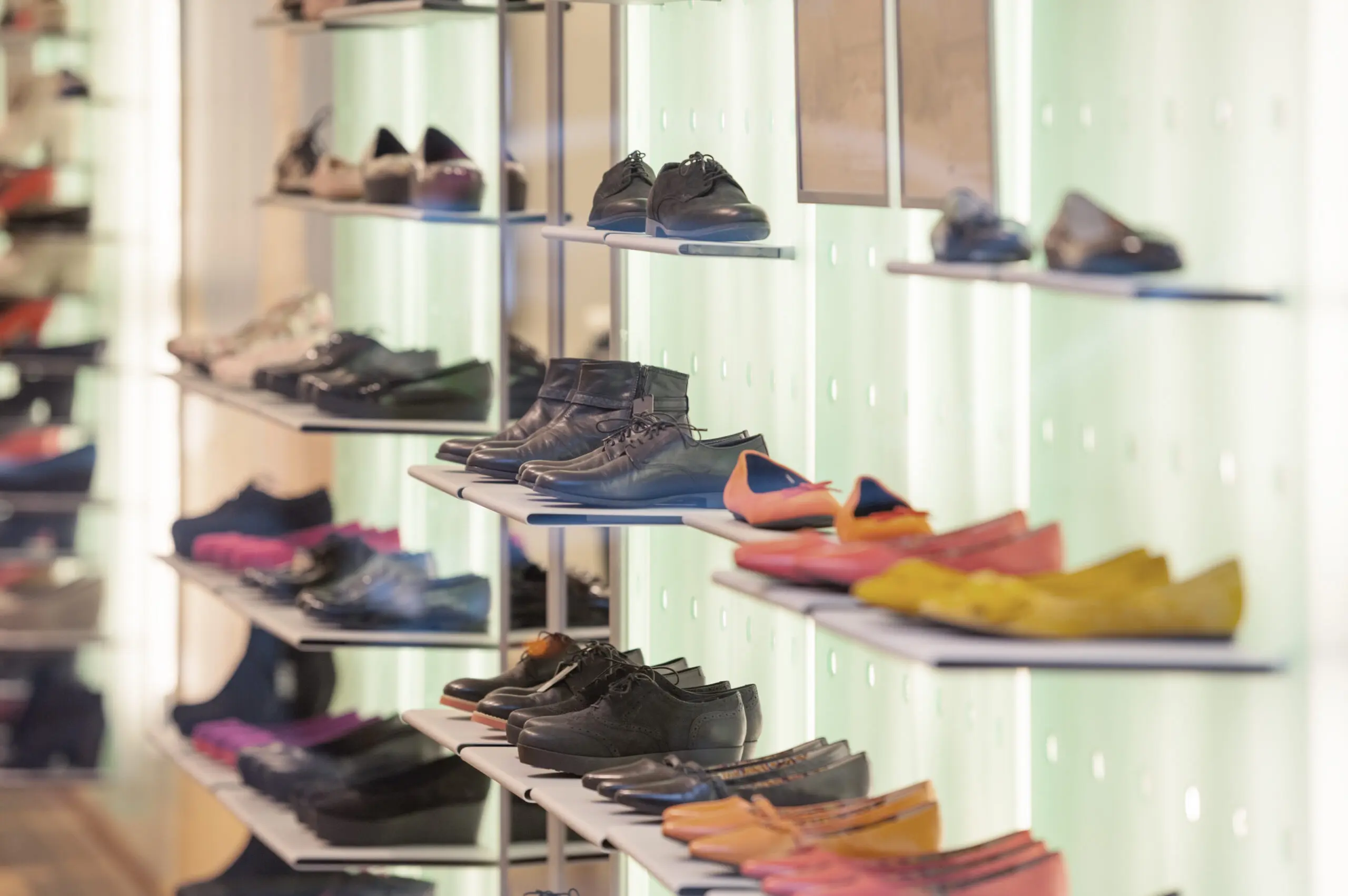 Adherent (formerly Compliance & Risks). A retail display of shoes on illuminated shelves, featuring men's dark leather shoes and boots, and women's colorful ballet flats in orange, pink, and yellow.