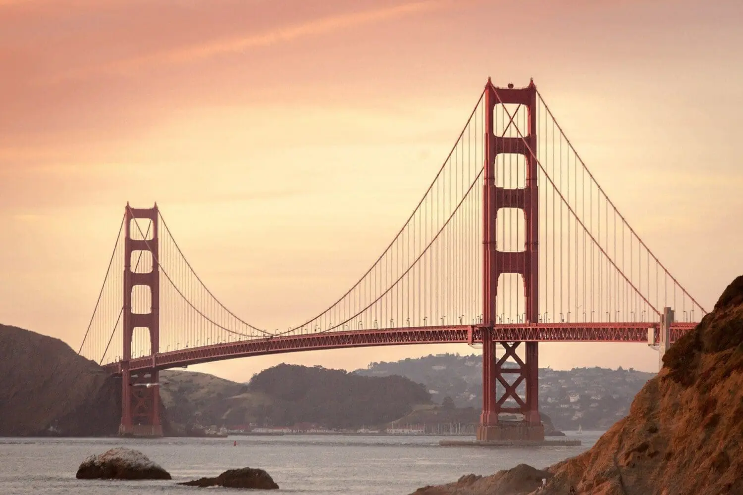 Adherent (formerly Compliance & Risks). The Golden Gate Bridge, a red-orange suspension bridge, spans across water under a soft orange and pink sky at sunrise or sunset. Rocky hills are visible on both sides.