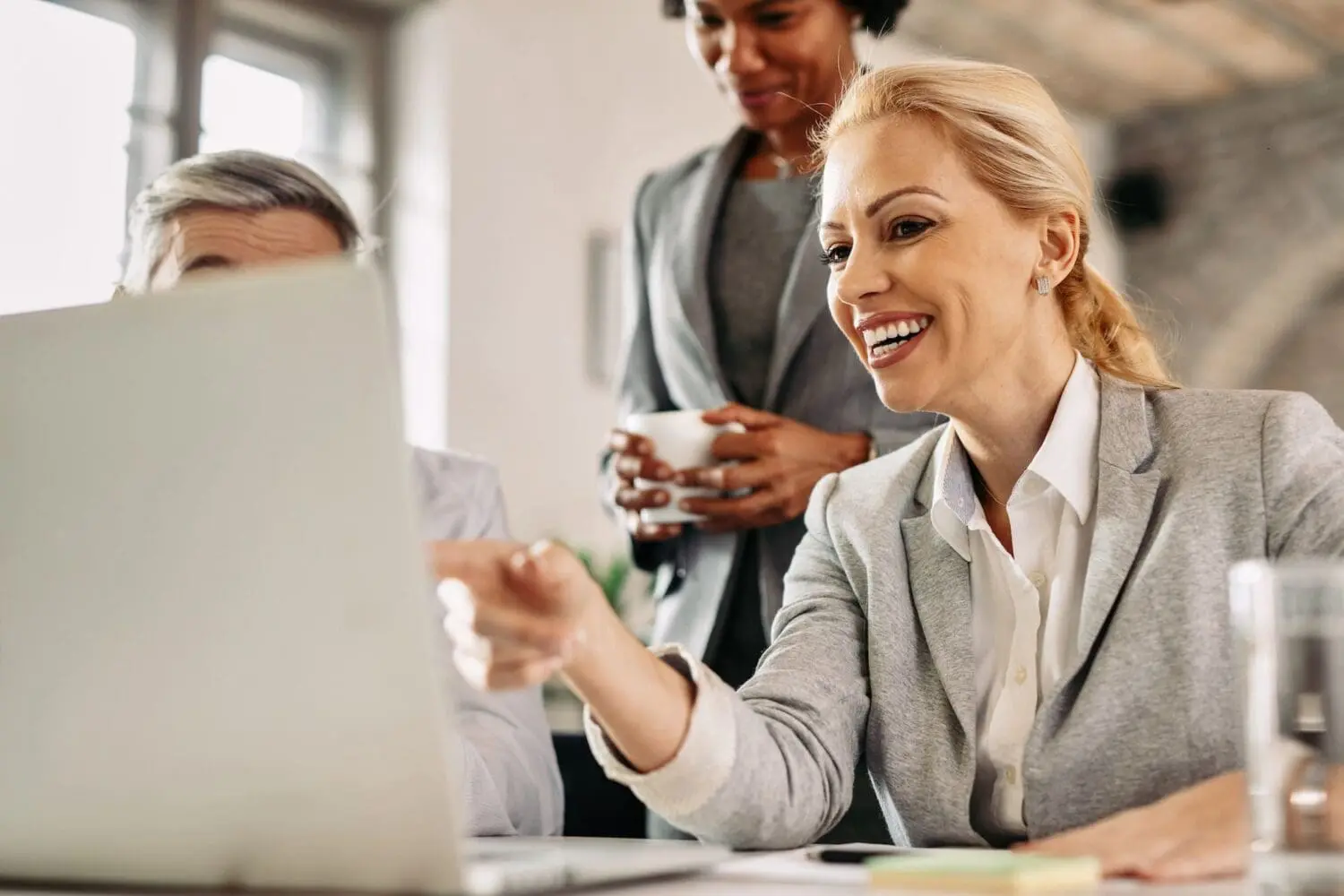 Adherent (formerly Compliance & Risks). Three business professionals, two women and one man, gather around a laptop in an office, with one woman pointing at the screen and smiling.