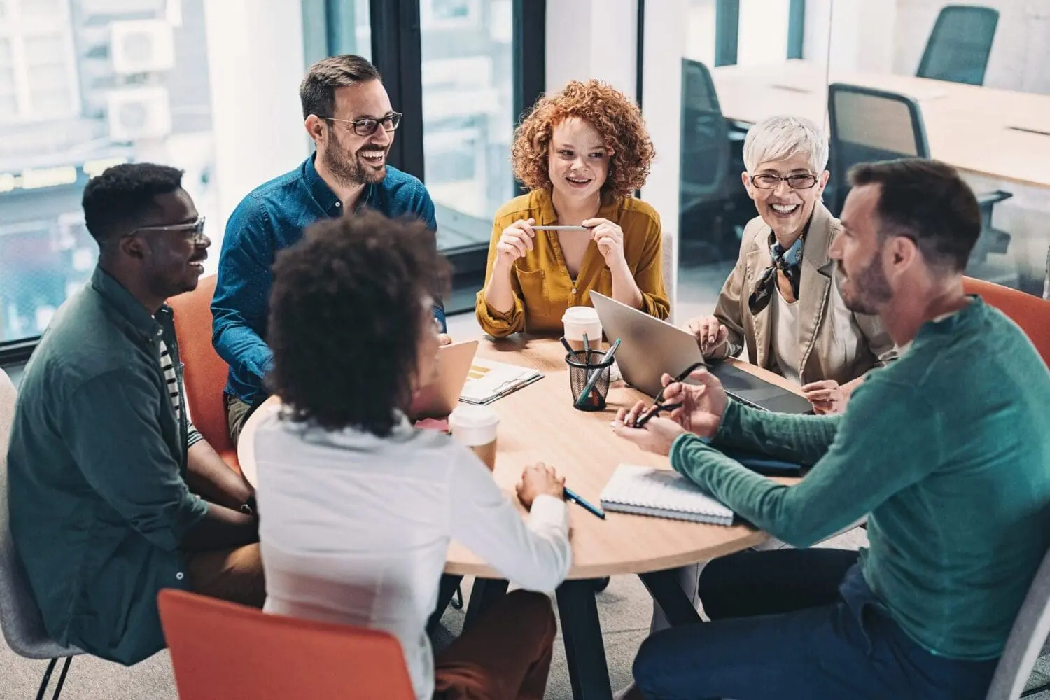 Adherent (formerly Compliance & Risks). Six diverse professionals are smiling and laughing while collaborating around a round table with laptops and coffee cups in a modern office.