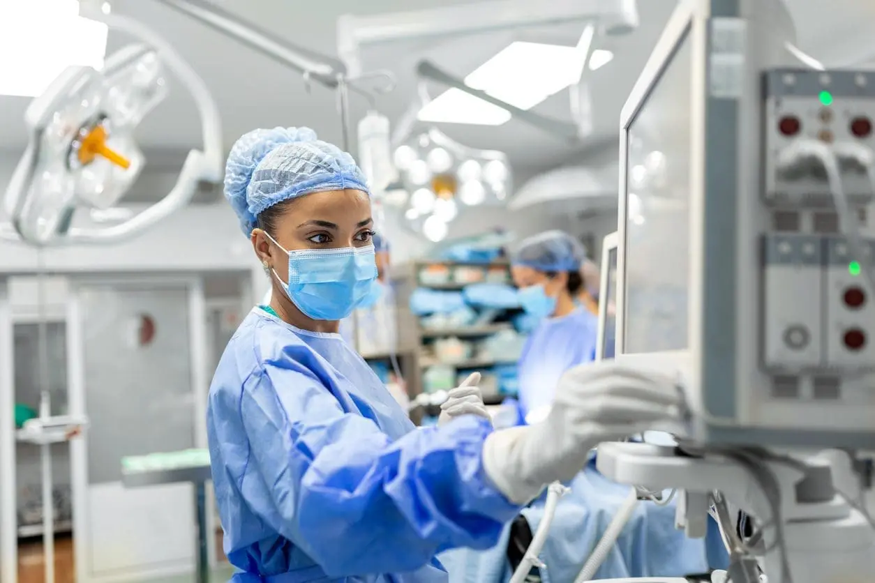 Adherent (formerly Compliance & Risks). A female medical professional in blue scrubs, mask, and gloves, operating equipment in a surgical room.