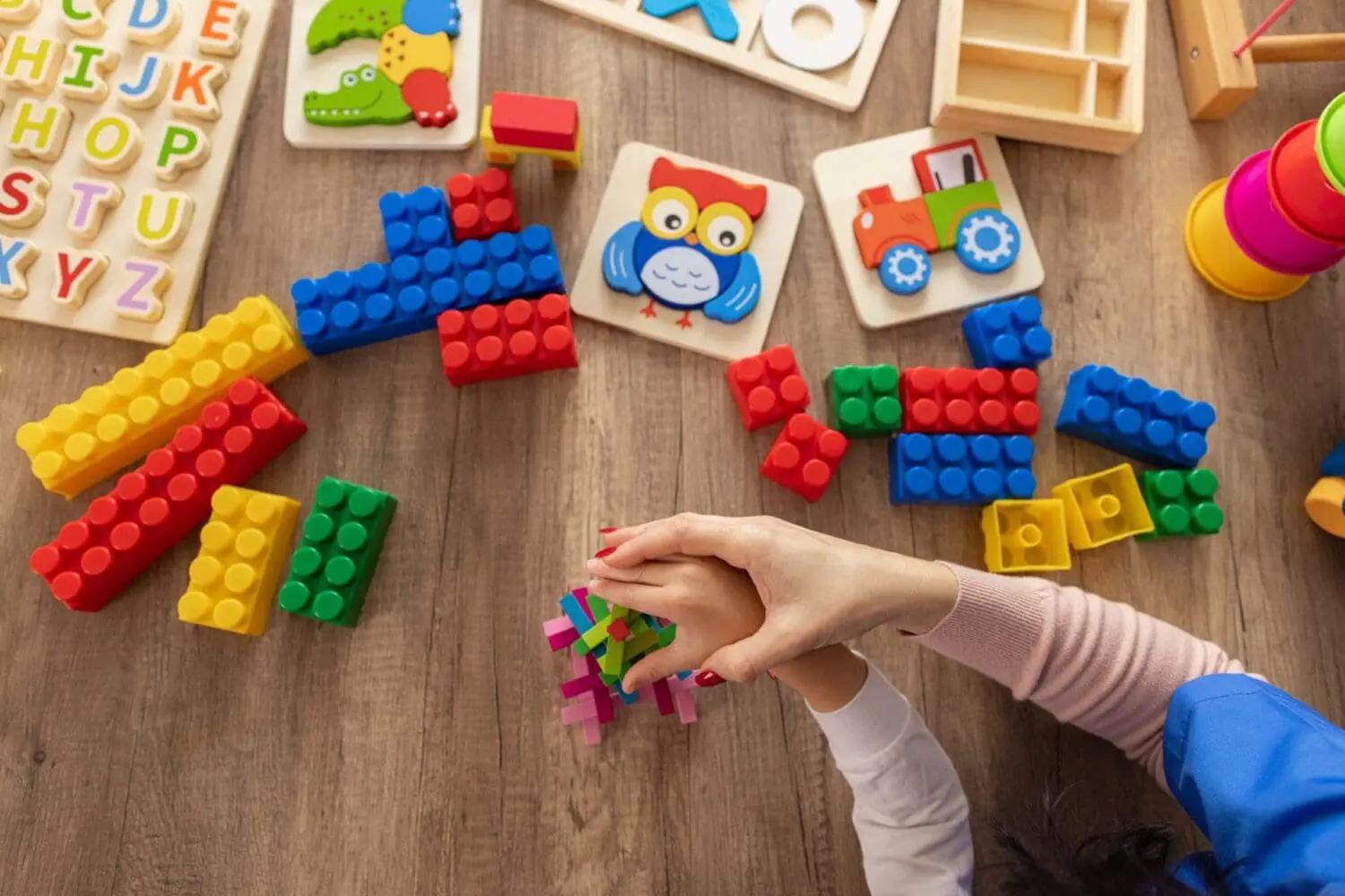 Overhead view of an adult and child's hands playing with colorful interlocking blocks on a wooden surface surrounded by various toys including building blocks, alphabet puzzles, and animal puzzles.