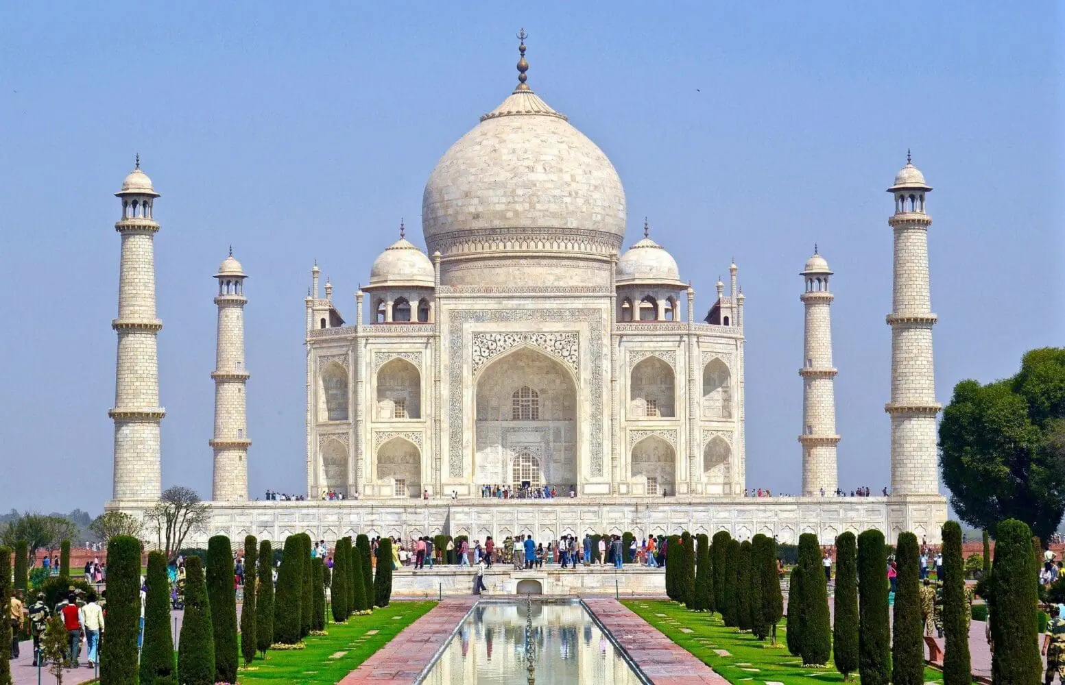 Adherent (formerly Compliance & Risks). The Taj Mahal, a white marble mausoleum with a large dome and four minarets, reflected in a long pool, surrounded by green lawns and conical trees under a clear blue sky.