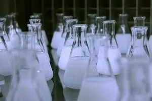 Adherent (formerly Compliance & Risks). Numerous clear glass Erlenmeyer flasks, each containing a white, opaque liquid, arranged in rows in a laboratory setting.