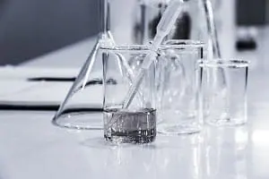 Adherent (formerly Compliance & Risks). Clear glass laboratory beakers and flasks on a light surface. One beaker contains two liquid layers and a glass dropper.