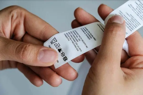 Adherent (formerly Compliance & Risks). Close-up of two hands holding a white clothing care label with laundry symbols and text.