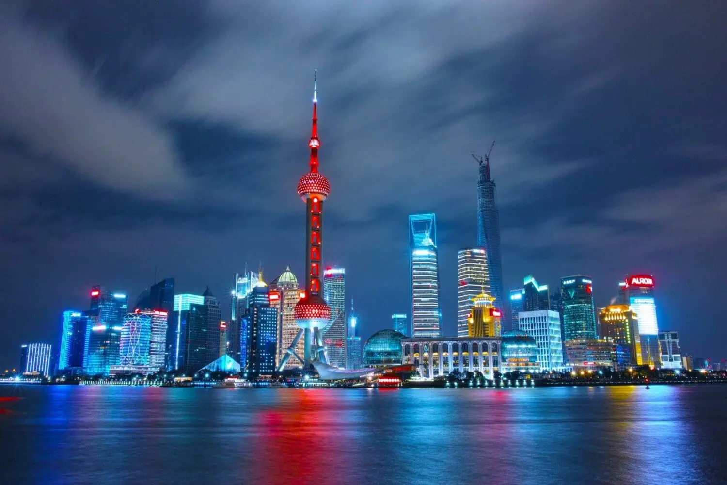 Nighttime view of the Shanghai skyline, featuring the red-lit Oriental Pearl TV Tower and other brightly illuminated skyscrapers reflecting on the water under a cloudy sky.