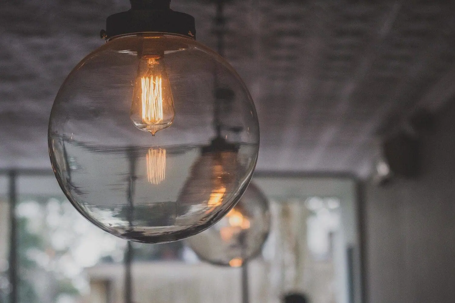 Adherent (formerly Compliance & Risks). A close-up of a clear spherical glass light fixture with a glowing Edison bulb inside, suspended from a ceiling. Another blurred light fixture is in the background.