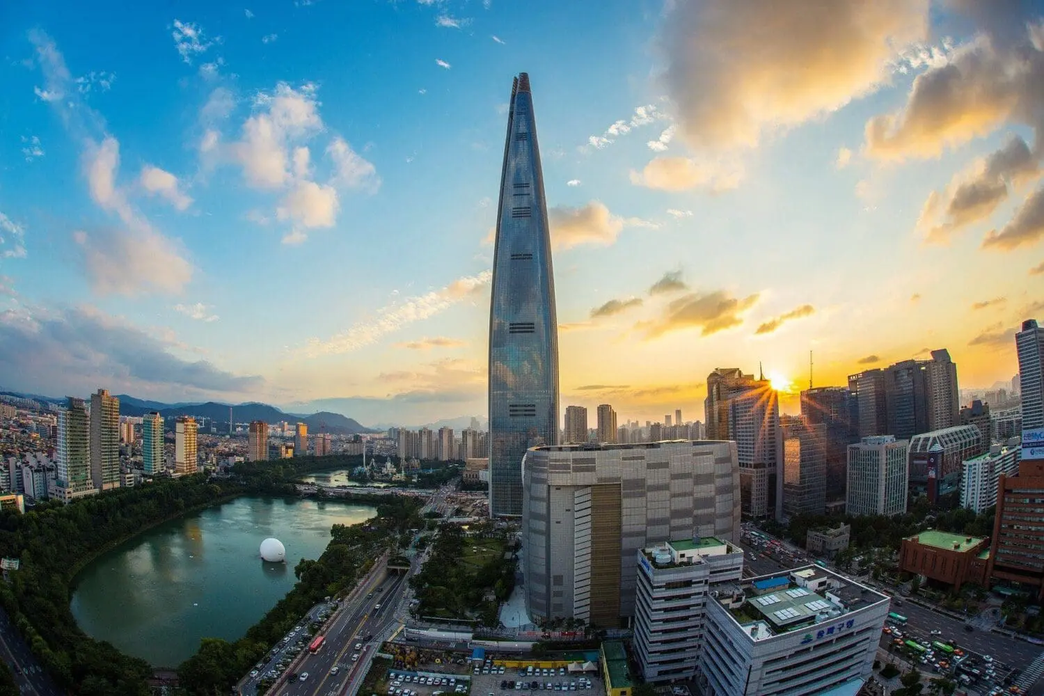Adherent (formerly Compliance & Risks). Aerial view of Seoul at sunset, featuring the towering Lotte World Tower, a large lake with a white sphere, and numerous city buildings under a colorful sky.