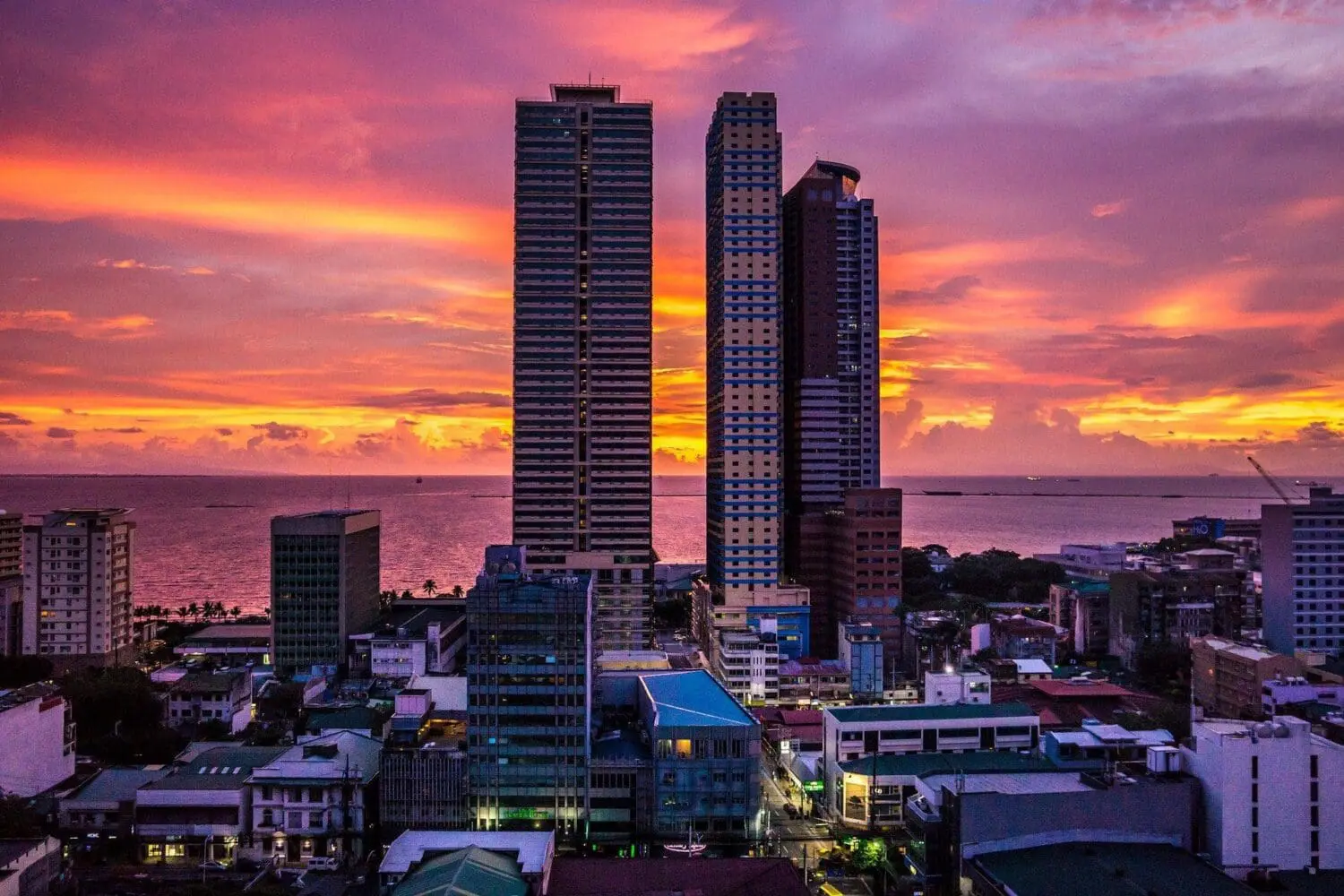 Adherent (formerly Compliance & Risks). Aerial view of a city at sunset with tall skyscrapers, smaller buildings, and a body of water under an orange, pink, and purple sky.