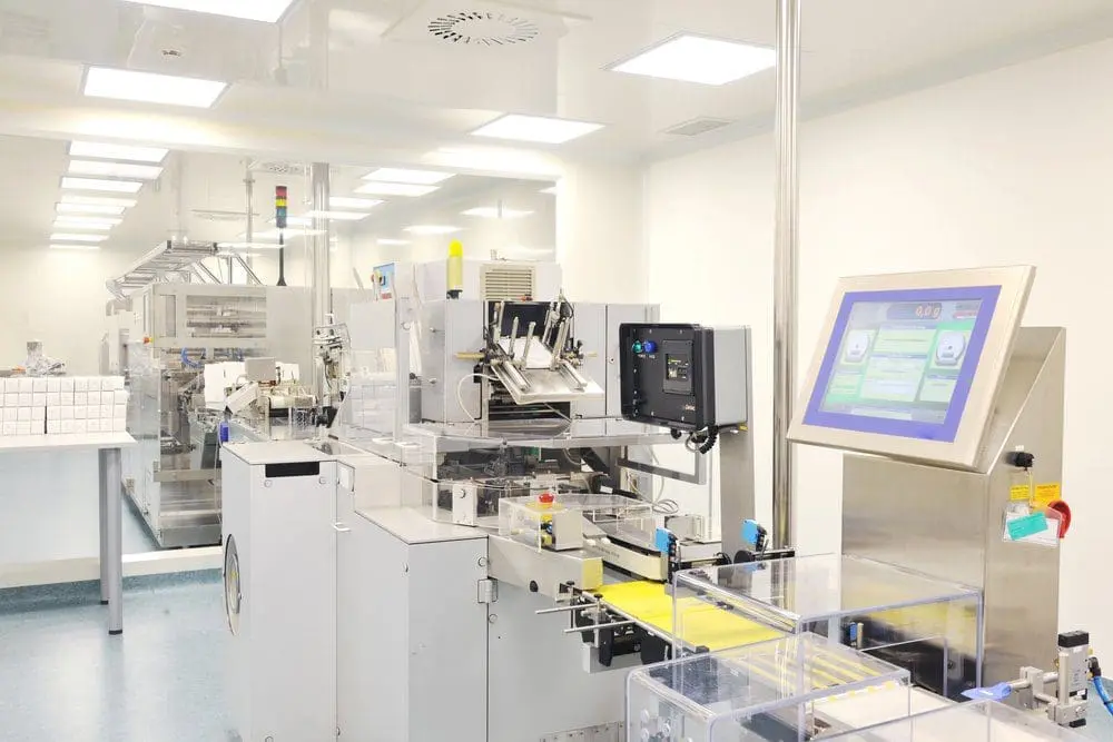 Adherent (formerly Compliance & Risks). Automated packaging machinery in a cleanroom, featuring conveyor belts, control screens, and stacked white boxes.