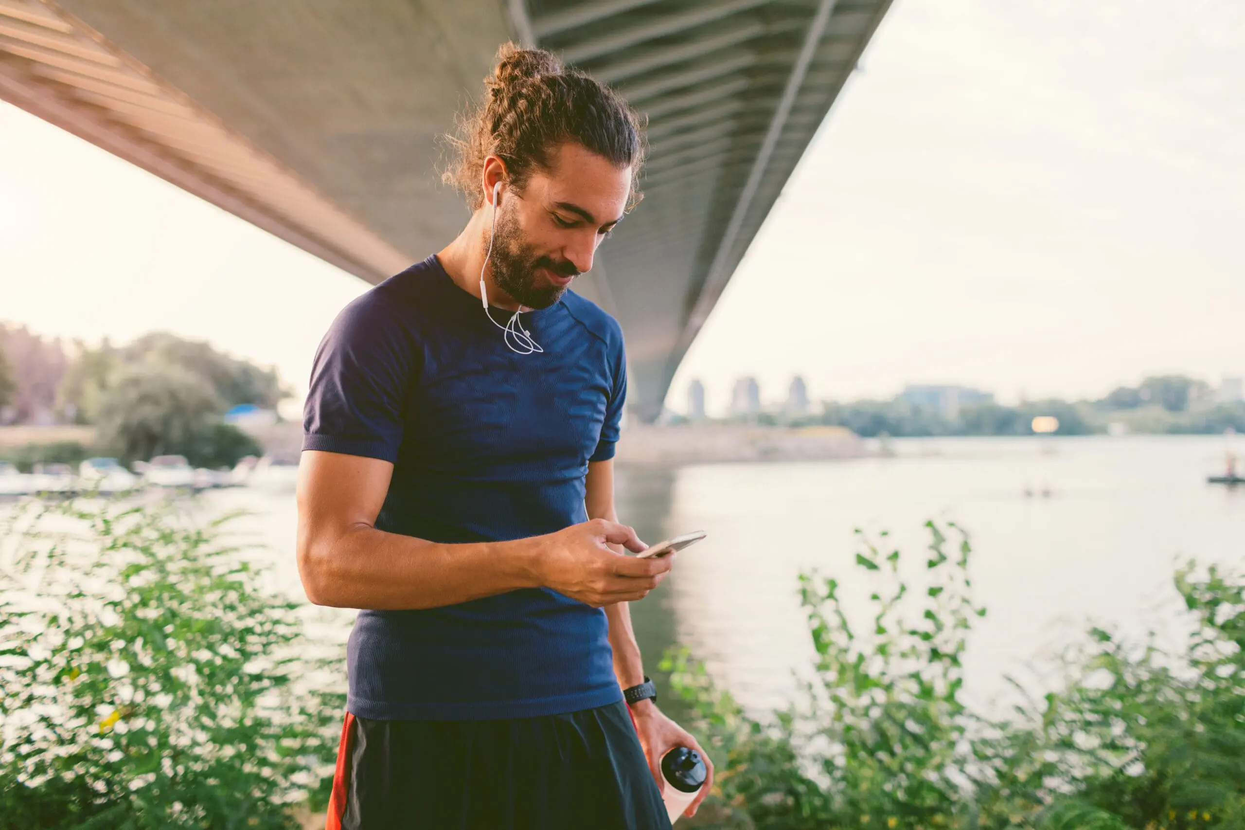 Adherent (formerly Compliance & Risks). A man with a man bun and beard, wearing athletic gear and earbuds, looks at his phone while holding a water bottle under a bridge.