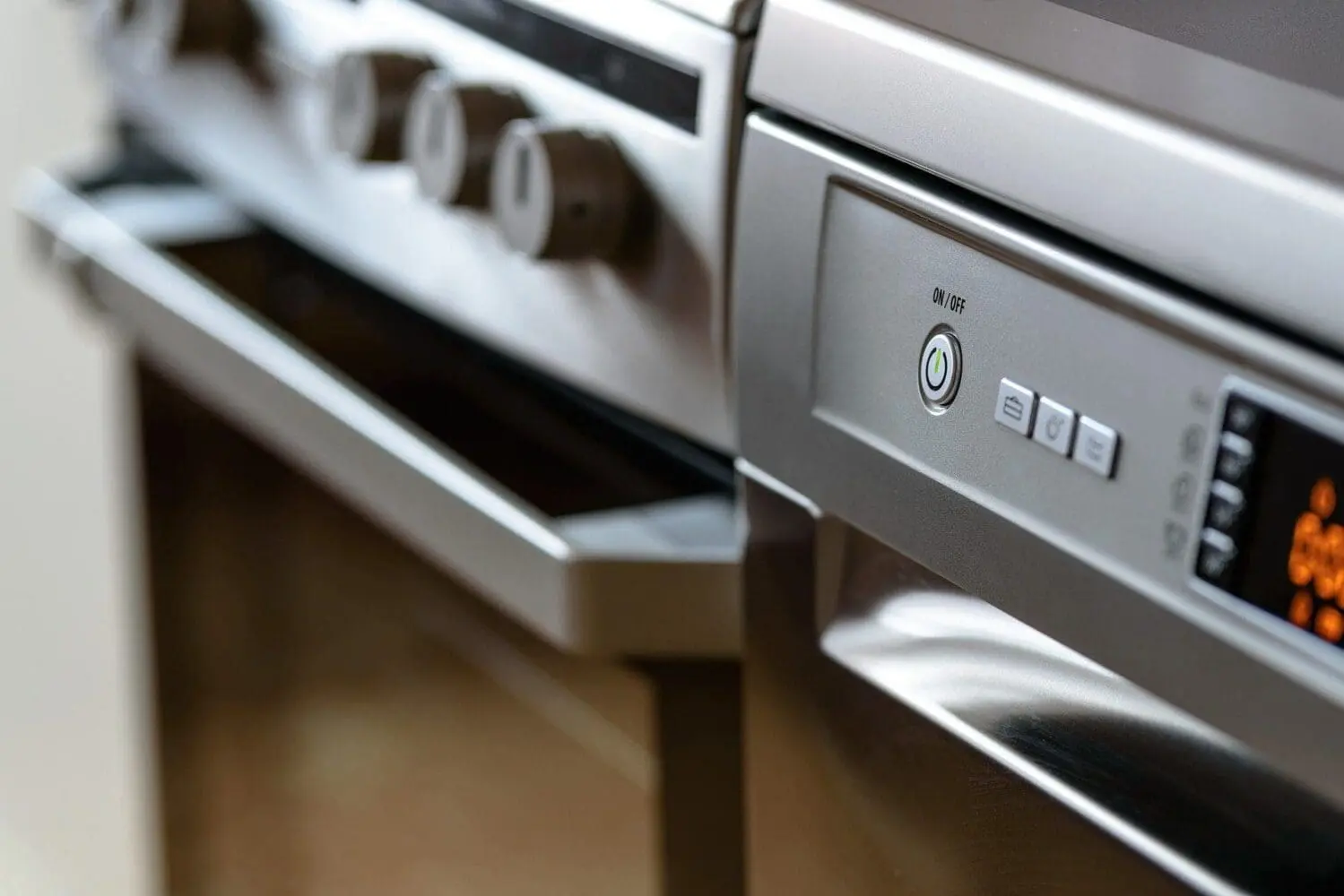 Close-up of a stainless steel dishwasher control panel with an illuminated green power button and digital display, next to a blurred oven.