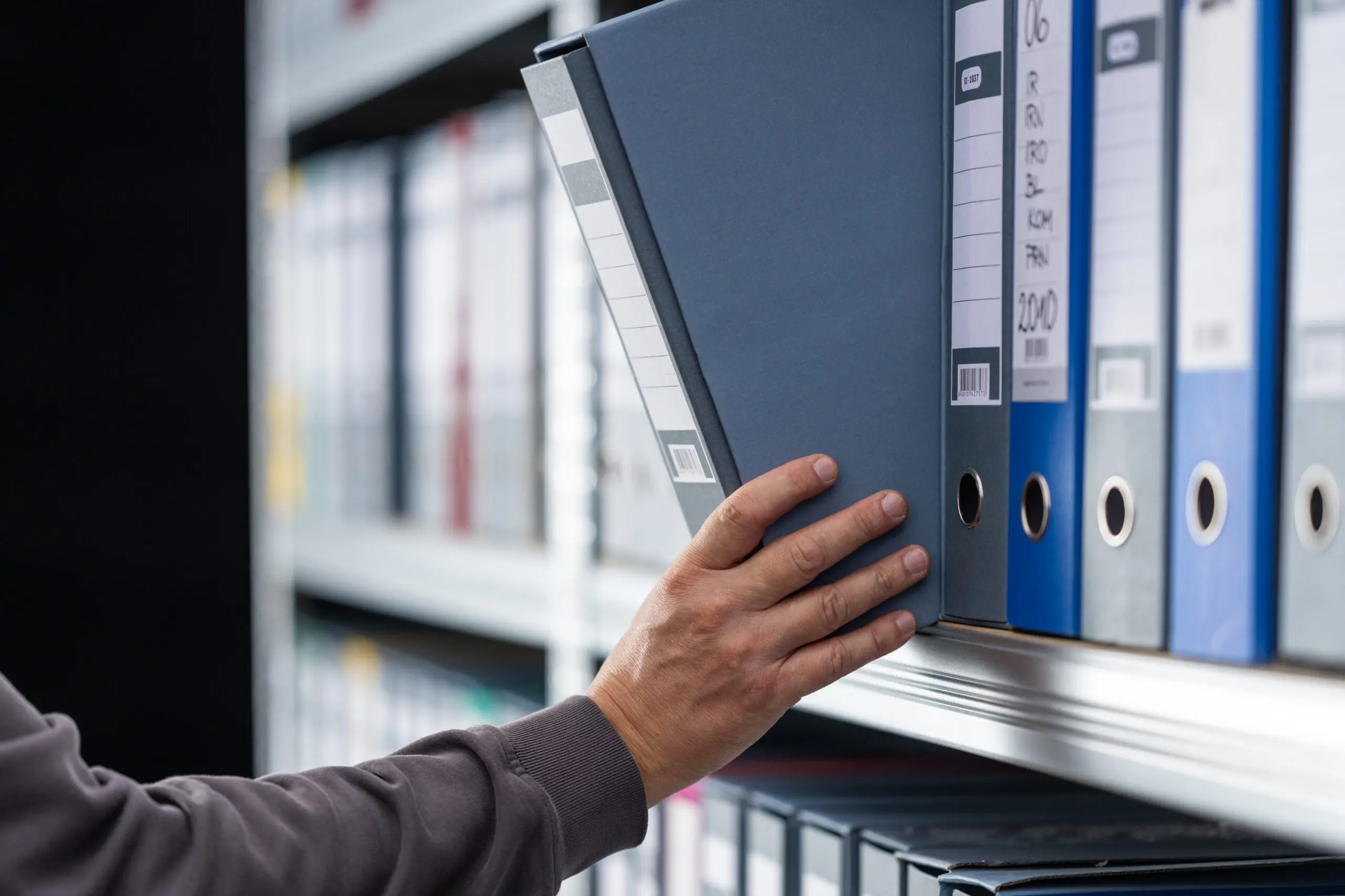 Adherent (formerly Compliance & Risks). A person's hand reaching for a dark blue binder on a shelf filled with various colored binders in an office setting.