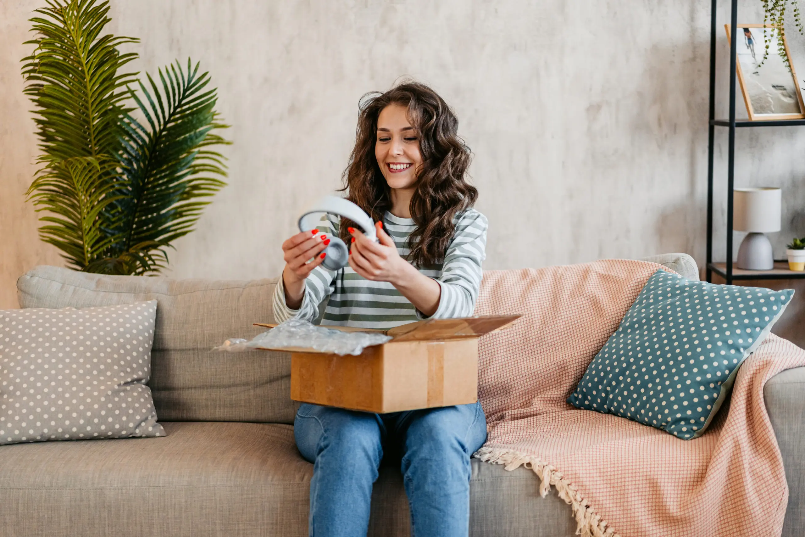 Adherent (formerly Compliance & Risks). A smiling woman unboxes light gray headphones from a cardboard box while sitting on a sofa in a living room.