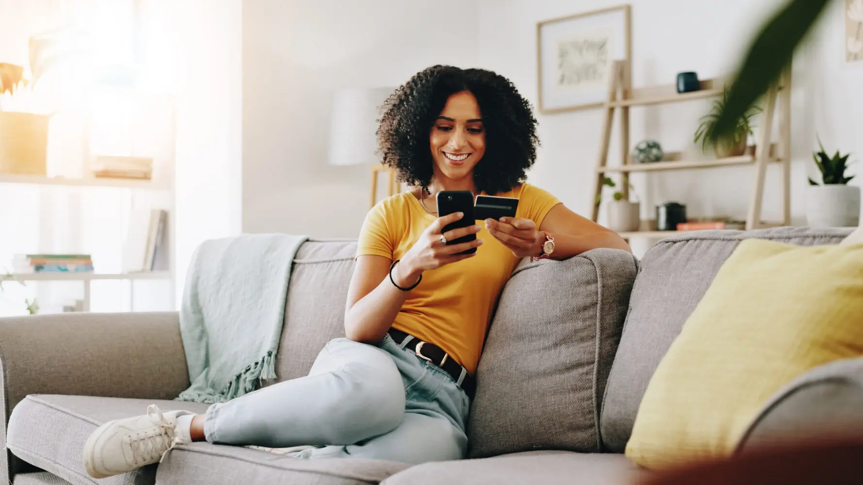 Adherent (formerly Compliance & Risks). A smiling woman with curly hair sits on a grey couch, holding a smartphone and a credit card, appearing to make an online purchase.