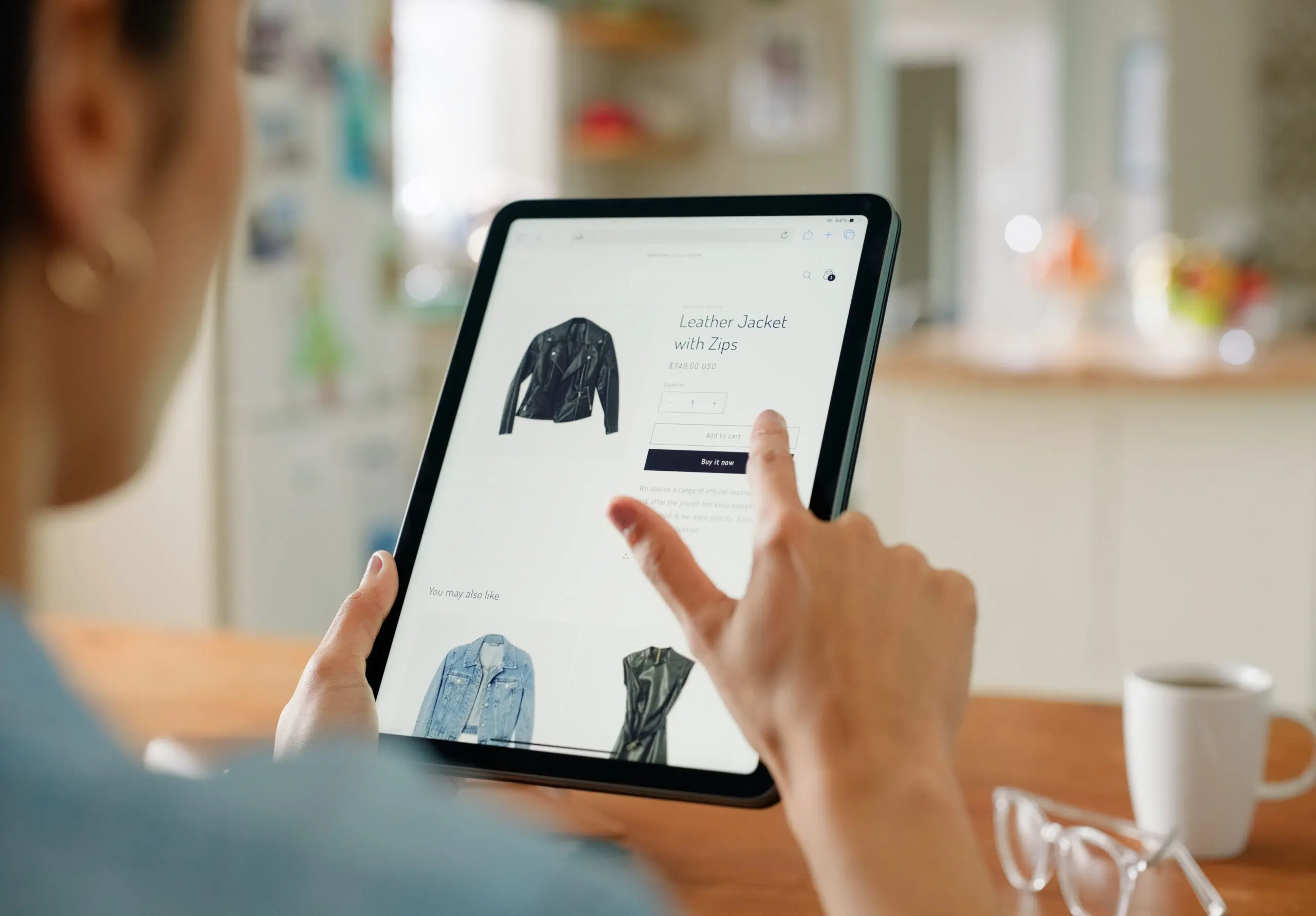 Adherent (formerly Compliance & Risks). A person's hands holding a tablet, displaying an e-commerce page for a black leather jacket with zips. The person's finger is touching the 'Buy it now' button.