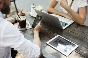 Adherent (formerly Compliance & Risks). Two people at a wooden table with a laptop, tablet, and smartphone, along with an iced coffee.