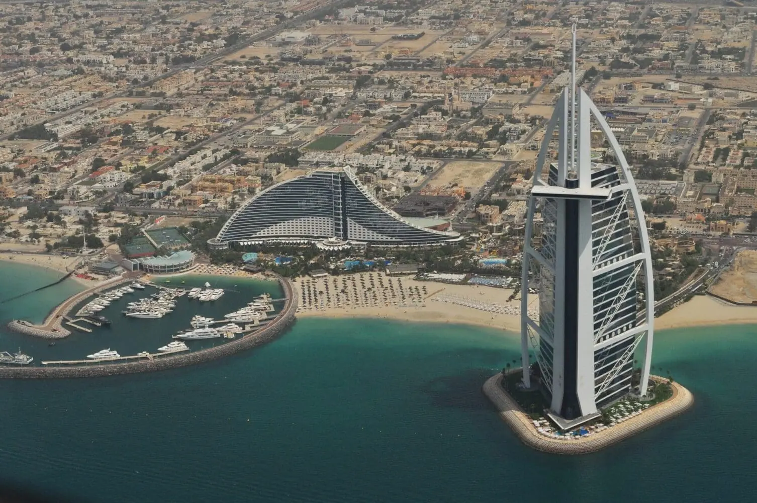 Adherent (formerly Compliance & Risks). Aerial view of Dubai showing the sail-shaped Burj Al Arab and wave-shaped Jumeirah Beach Hotel, a marina with yachts, sandy beaches, and the city skyline.