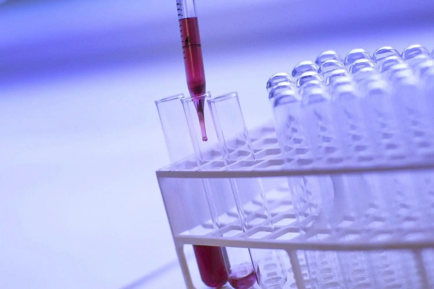Adherent (formerly Compliance & Risks). A close-up of a pipette dispensing red liquid into a clear test tube in a white rack, with other test tubes visible.