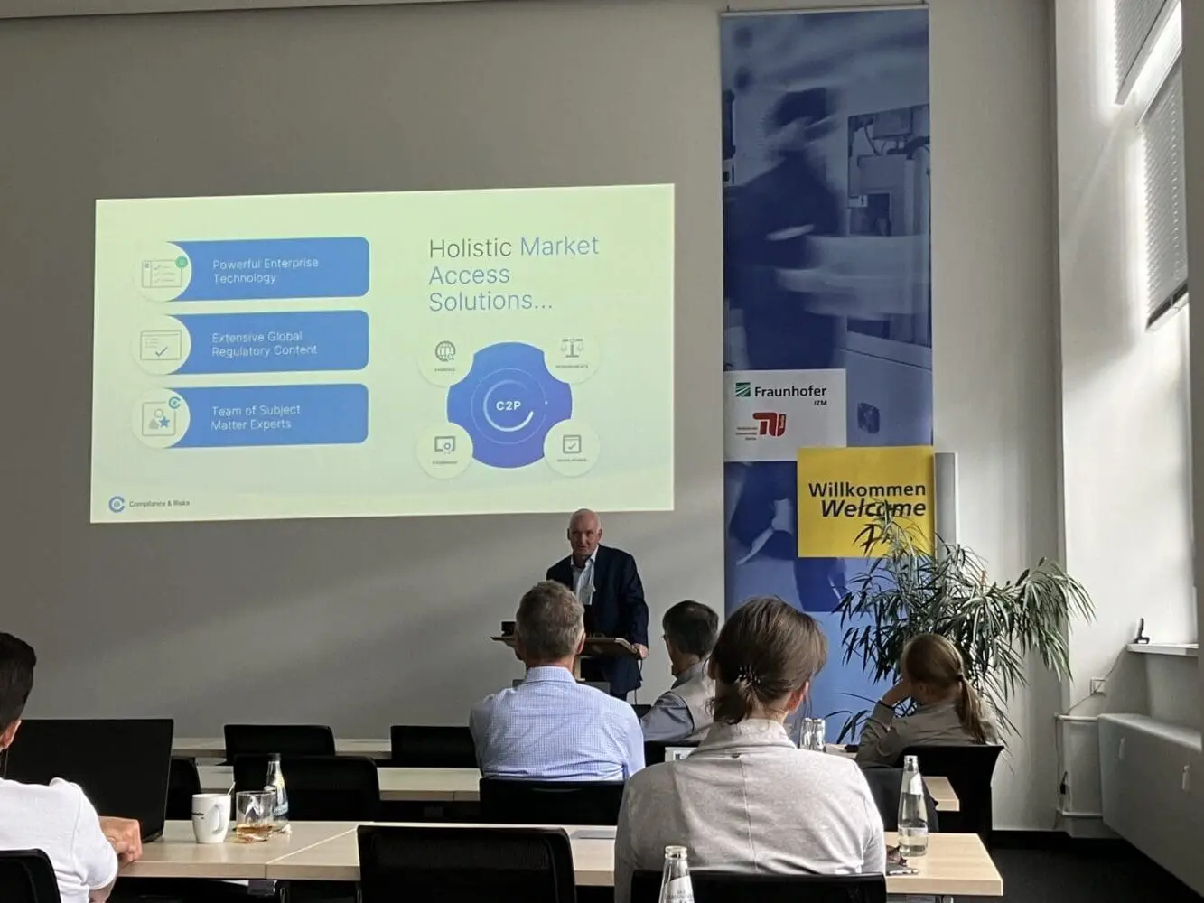 Adherent (formerly Compliance & Risks). A man presents a slide titled 'Holistic Market Access Solutions' to an audience in a conference room. The slide mentions 'Powerful Enterprise Technology' and 'Extensive Global Regulatory Content'. Banners for Fraunhofer IZM and a 'Welcome' sign are visible in the background.