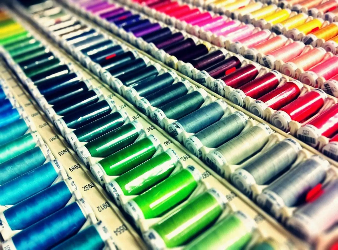 Adherent (formerly Compliance & Risks). Close-up of numerous spools of colorful sewing thread arranged in rows, showing a gradient of hues from blues and greens to reds, pinks, purples, and yellows.
