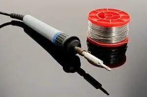 Adherent (formerly Compliance & Risks). A soldering iron with a gray handle and a metallic tip next to a red spool of silver solder wire, both on a dark reflective surface.