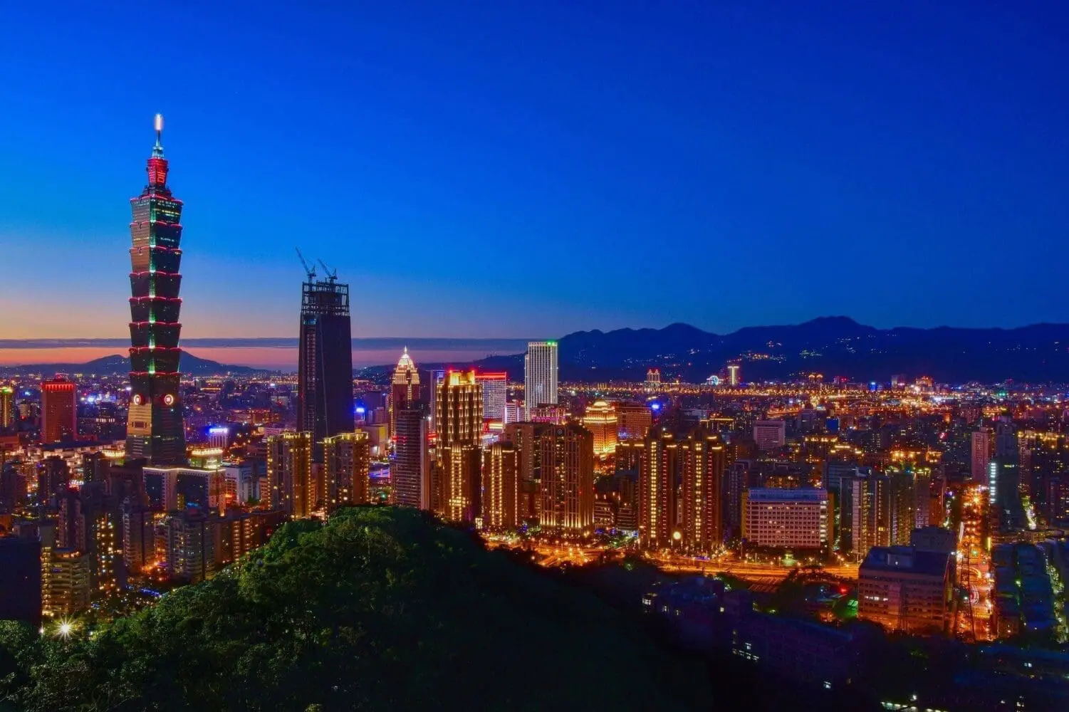 Adherent (formerly Compliance & Risks). Panoramic view of Taipei city skyline at twilight, featuring the illuminated Taipei 101 skyscraper, other glowing buildings, and mountains in the background.