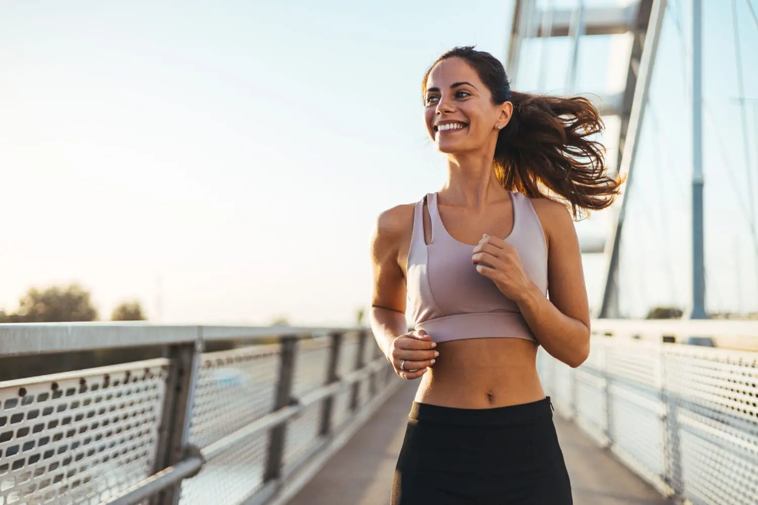 Adherent (formerly Compliance & Risks). A smiling woman with dark hair in a ponytail runs on a bridge, wearing a pink sports bra and black shorts, under a bright sky.