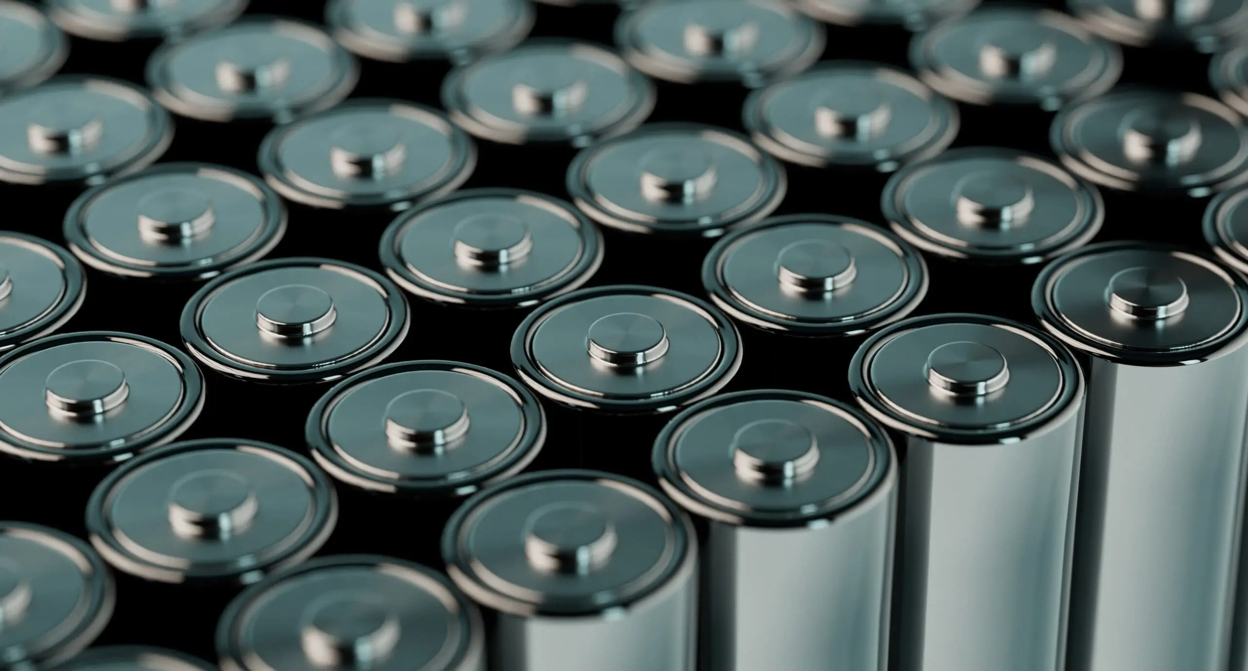 A close-up view of many silver cylindrical batteries with metallic tops arranged in rows on a dark background.