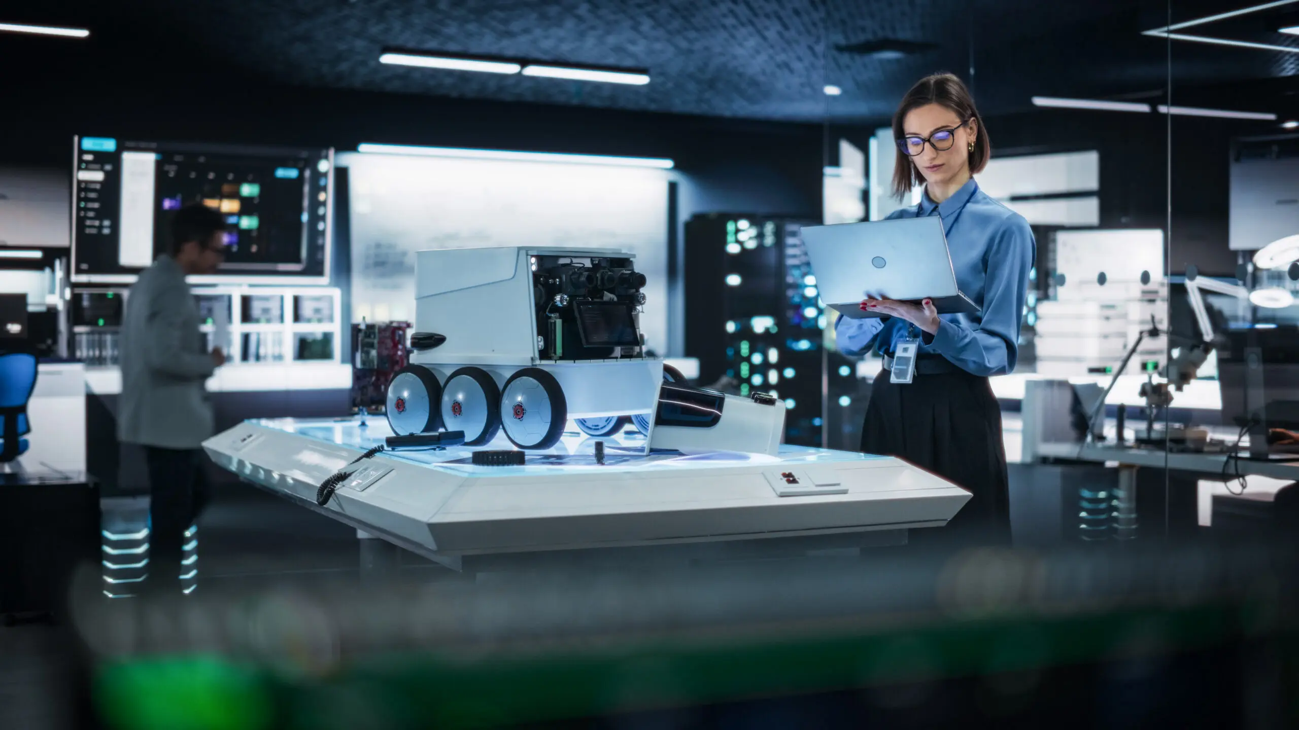 Adherent (formerly Compliance & Risks). A woman with glasses and a blue shirt holds a laptop next to a white, six-wheeled robot on an illuminated platform in a high-tech lab.