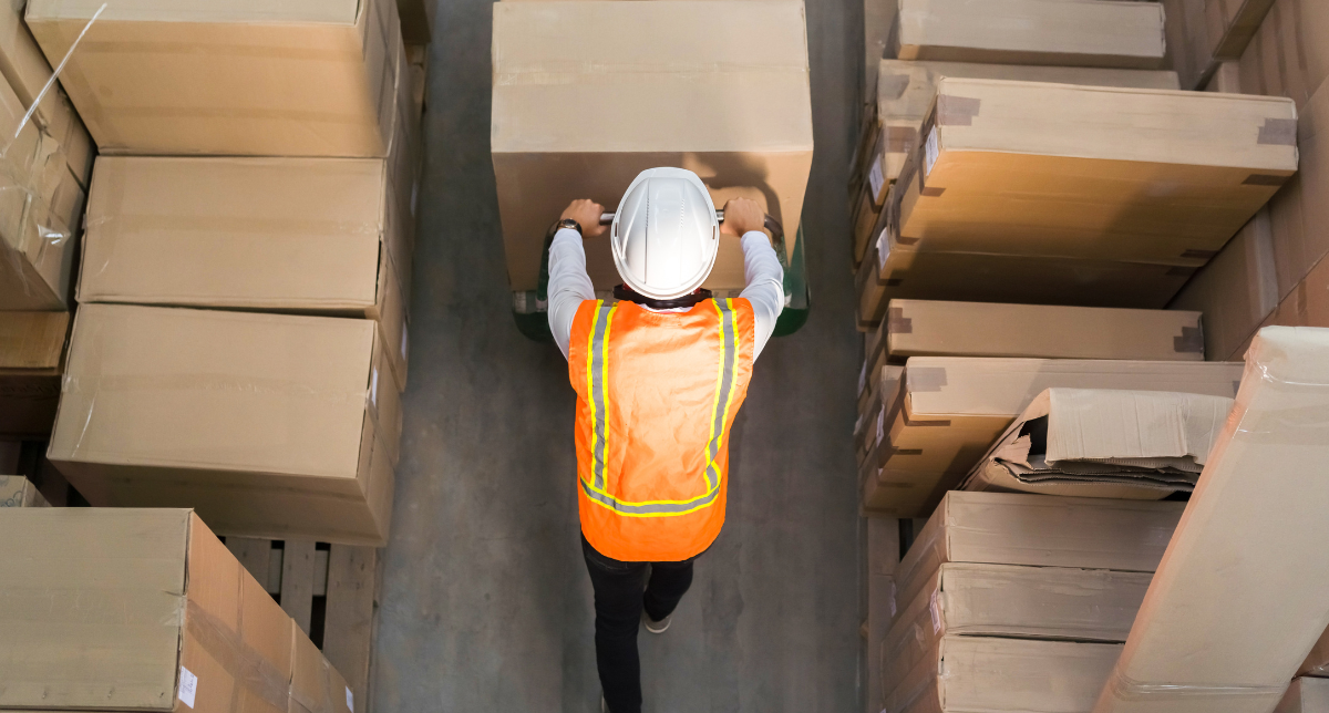 Overhead view of a warehouse worker in a hard hat and orange safety vest pushing a large cardboard box on a hand truck down an aisle lined with stacked boxes.