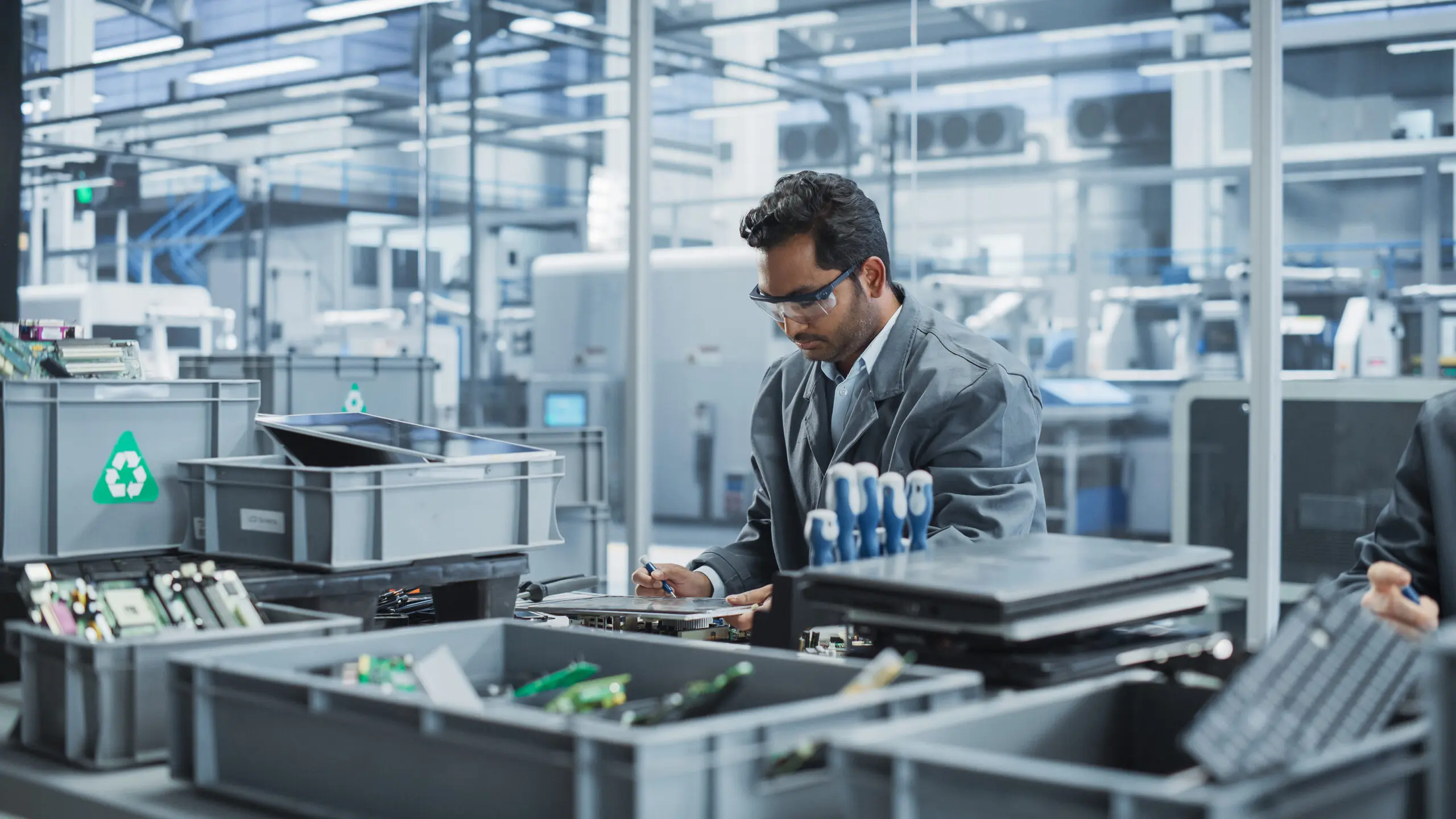 Adherent (formerly Compliance & Risks). A man in safety glasses and a grey jacket inspects electronic components on a table, surrounded by bins of e-waste in an industrial setting.