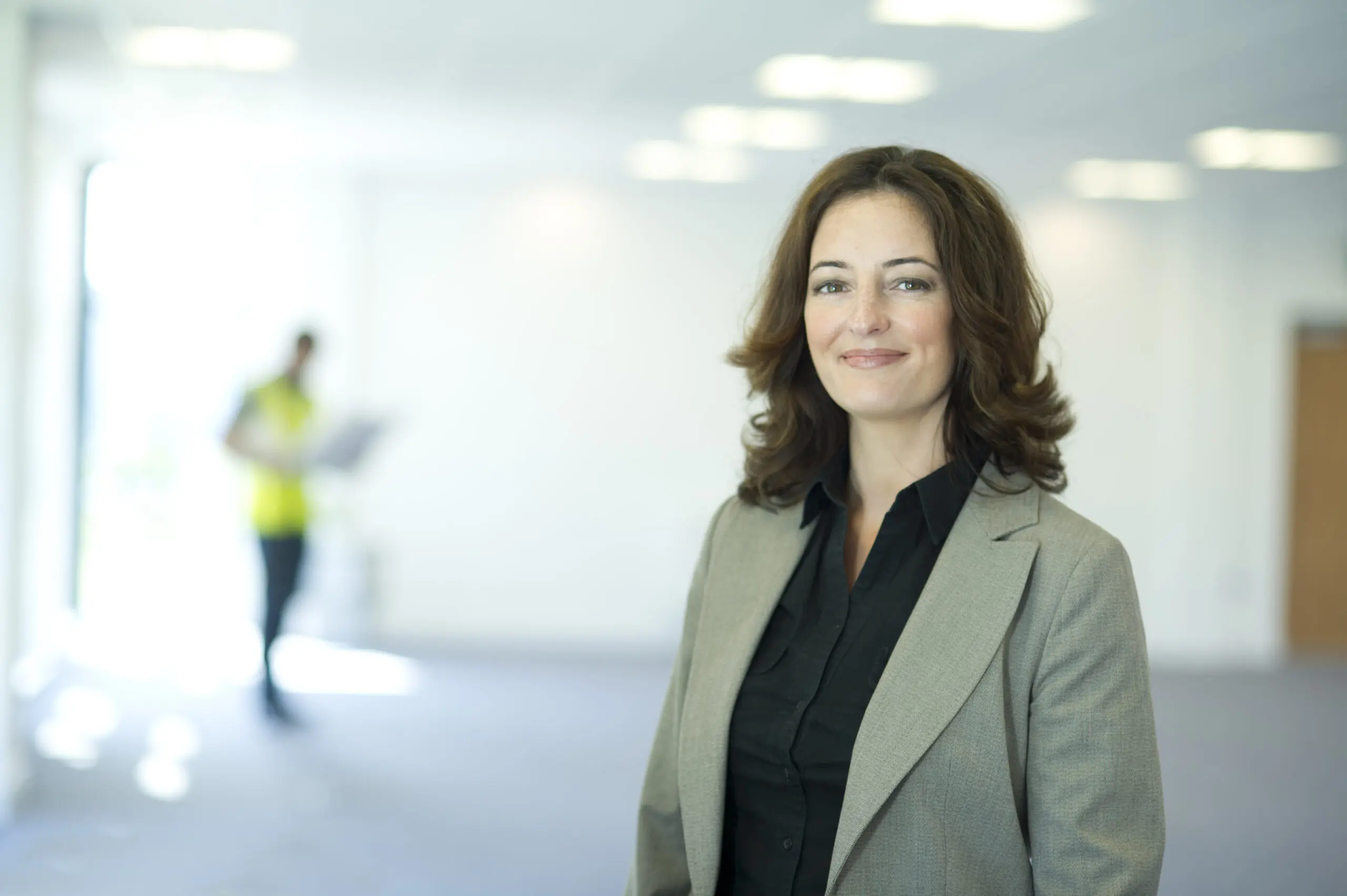 Adherent (formerly Compliance & Risks). A smiling woman in a grey blazer and black shirt stands in a bright, empty office with a blurred figure in a yellow vest in the background.