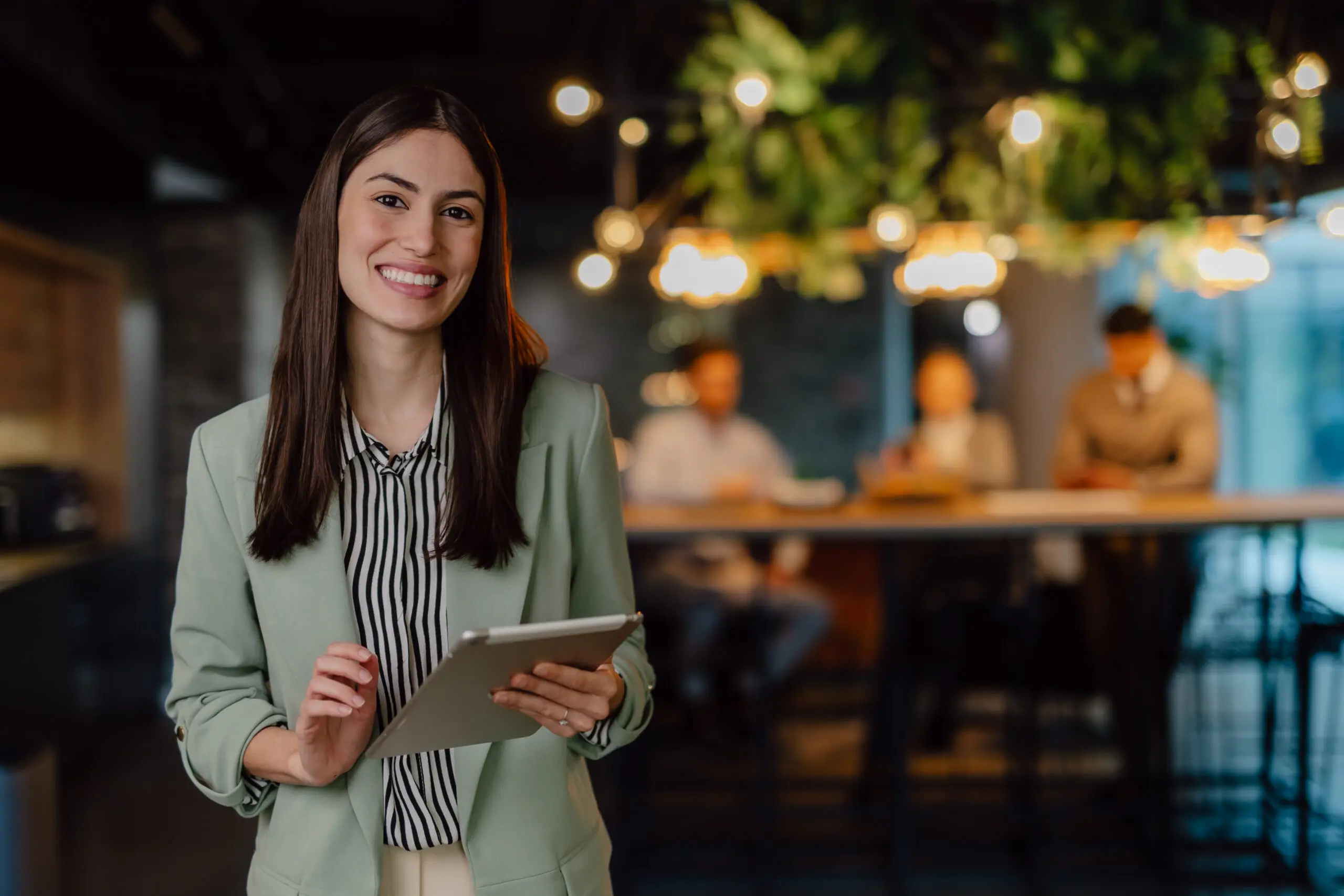 Adherent (formerly Compliance & Risks). A smiling young woman in a green blazer holds a tablet in a blurred office setting with warm lights and foliage.
